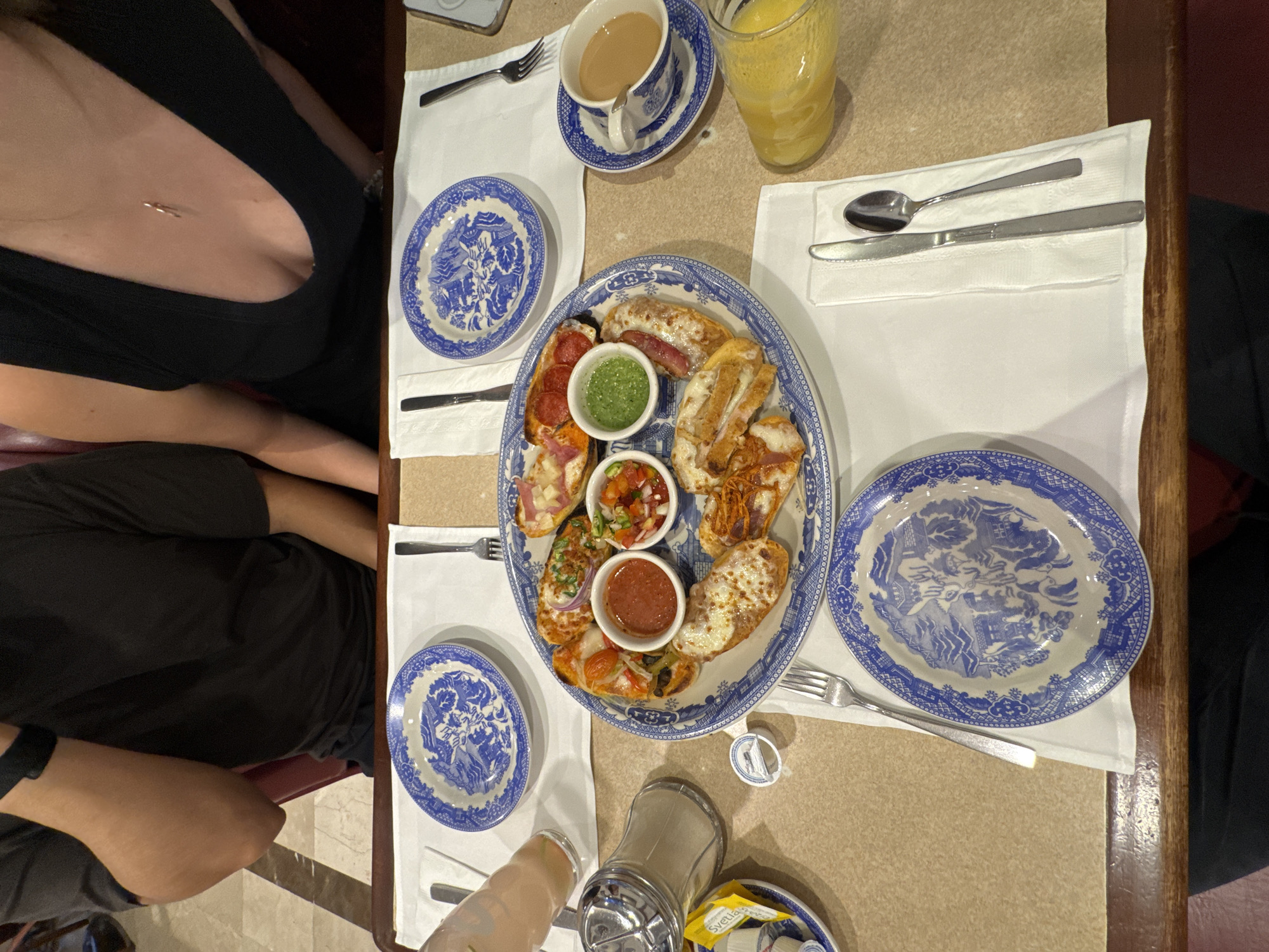 A top-down indoor shot shows a table with a light brown surface. At least two people are seated at the table, one wearing a black top with a low neckline and another wearing a black shirt. The table is set with white napkins and blue and white patterned china plates. A large oval plate contains a variety of open-faced sandwiches and small bowls of sauces. Other items on the table include a cup of tea, a glass of orange juice, silverware, a sugar dispenser, and small packets of sweetener. The scene is likely set in a restaurant or dining establishment in Puebla, Mexico.