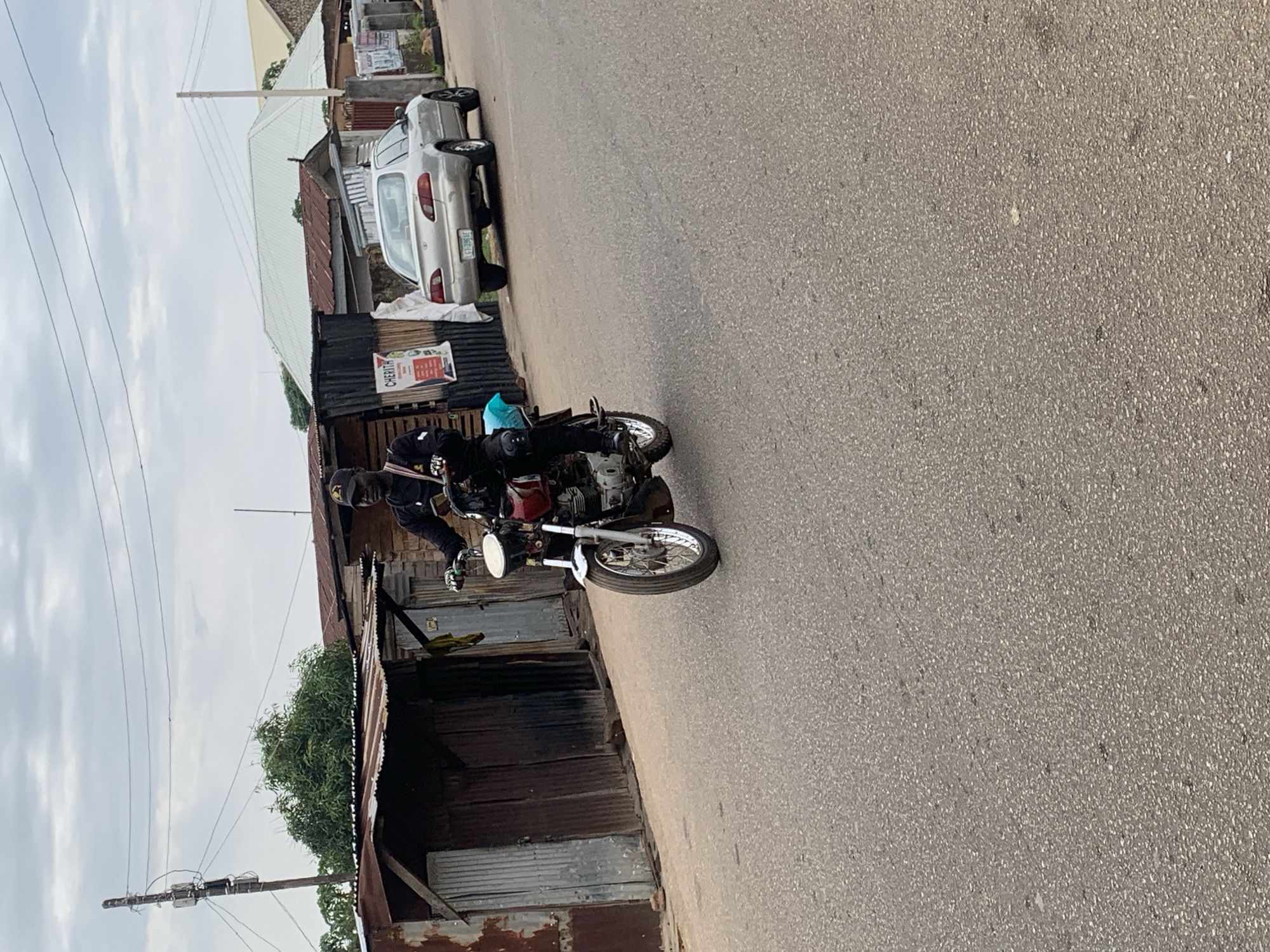 The image captures an outdoor street scene in Jos, Nigeria, under an overcast sky. In the central foreground, a dark-skinned male, wearing a black long-sleeved top, black trousers, black cap with a gold emblem, and dark gloves, is actively riding a motorcycle. The motorcycle features a dark red fuel tank, silver handlebars, and a white headlight. A light blue-wrapped item is secured on the rear seat of the motorcycle.
On the left side of the frame, behind the motorcyclist, are several small, single-story structures constructed from weathered and rusty corrugated metal sheets, likely serving as shops or dwellings. A wooden utility pole with multiple wires and attachments stands prominently on the far left. Green foliage is visible behind these structures.
Further down the street on the right side, a silver sedan, consistent with a Toyota Corolla, is parked facing away from the viewer. Its rear license plate, "LGT-396 AE," is clearly visible. Behind the parked car, additional buildings with various roof types, including a larger structure with a light-colored gabled roof and others with reddish-brown corrugated roofs, are present. A vertical signpost with some text, including "CHERITH" and "AGAIN," is partially visible near the car.
The road surface is asphalt, appearing dry and somewhat textured. The overall environment suggests a bustling, semi-urban or commercial area.