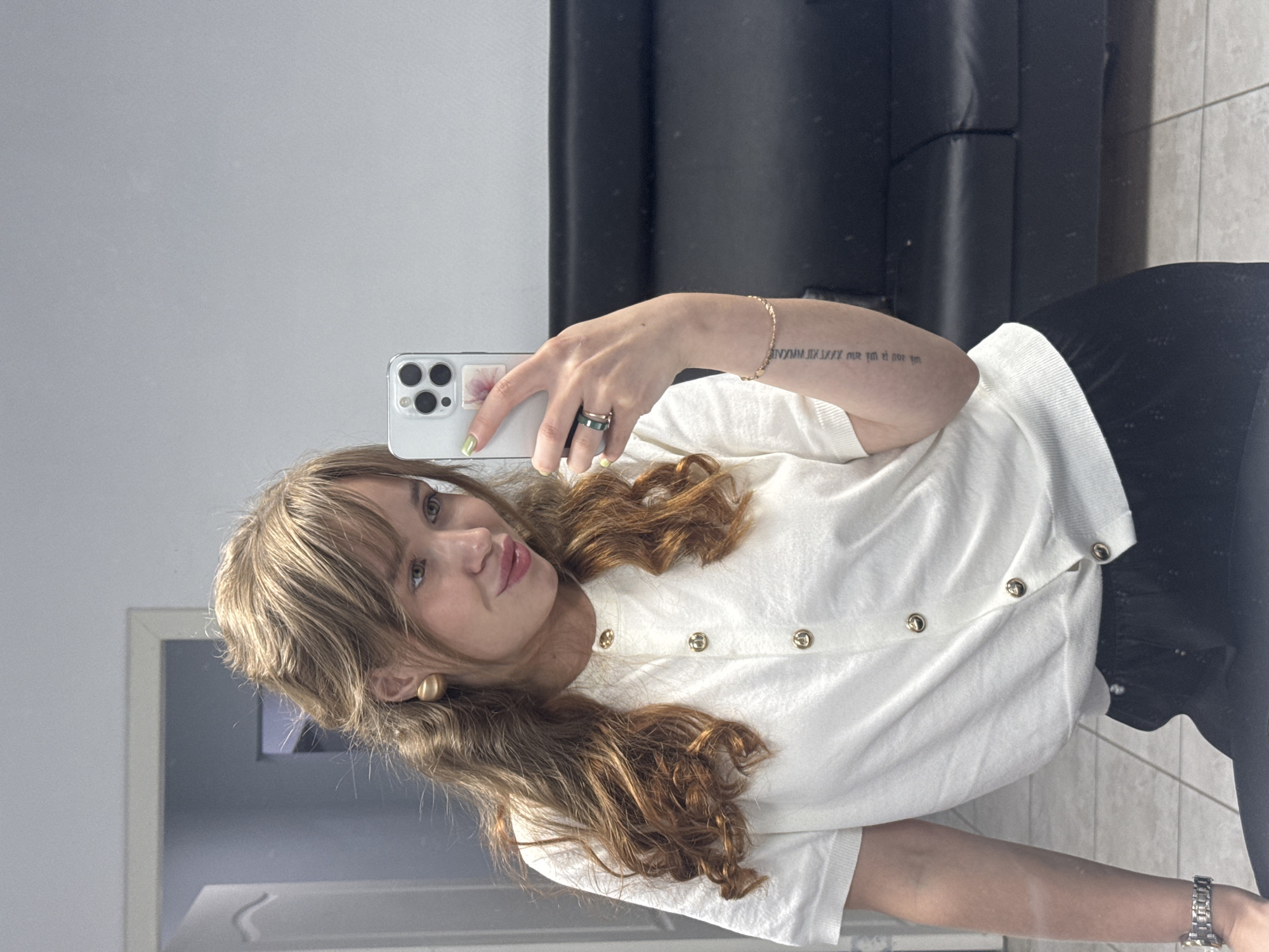 An indoor mirror selfie taken in Chaikovsky, Russia, features a young woman with long, wavy, reddish-brown hair and bangs. She is seen from the chest up, wearing a white short-sleeved top adorned with gold-colored buttons and black bottoms. She sports large, round, gold-colored earrings.
Her left hand holds a light-colored smartphone with multiple camera lenses and a small floral sticker on the back, angled to capture her reflection. Her fingernails are painted with light green polish. On her left hand, she wears a prominent dark ring and a thinner gold-colored ring. Her right forearm displays a delicate gold-colored chain bracelet and a silver watch on her wrist. A tattoo is visible on her right forearm, bearing the text "XXMM.II.XXXX nur für mich".
The woman exhibits a calm expression with a subtle smile. The background reflects a simple indoor setting, including a light grey wall, a dark black couch or piece of furniture, and a light-tiled floor. A doorframe or another reflective surface is partially visible on the left side of the mirror's reflection.