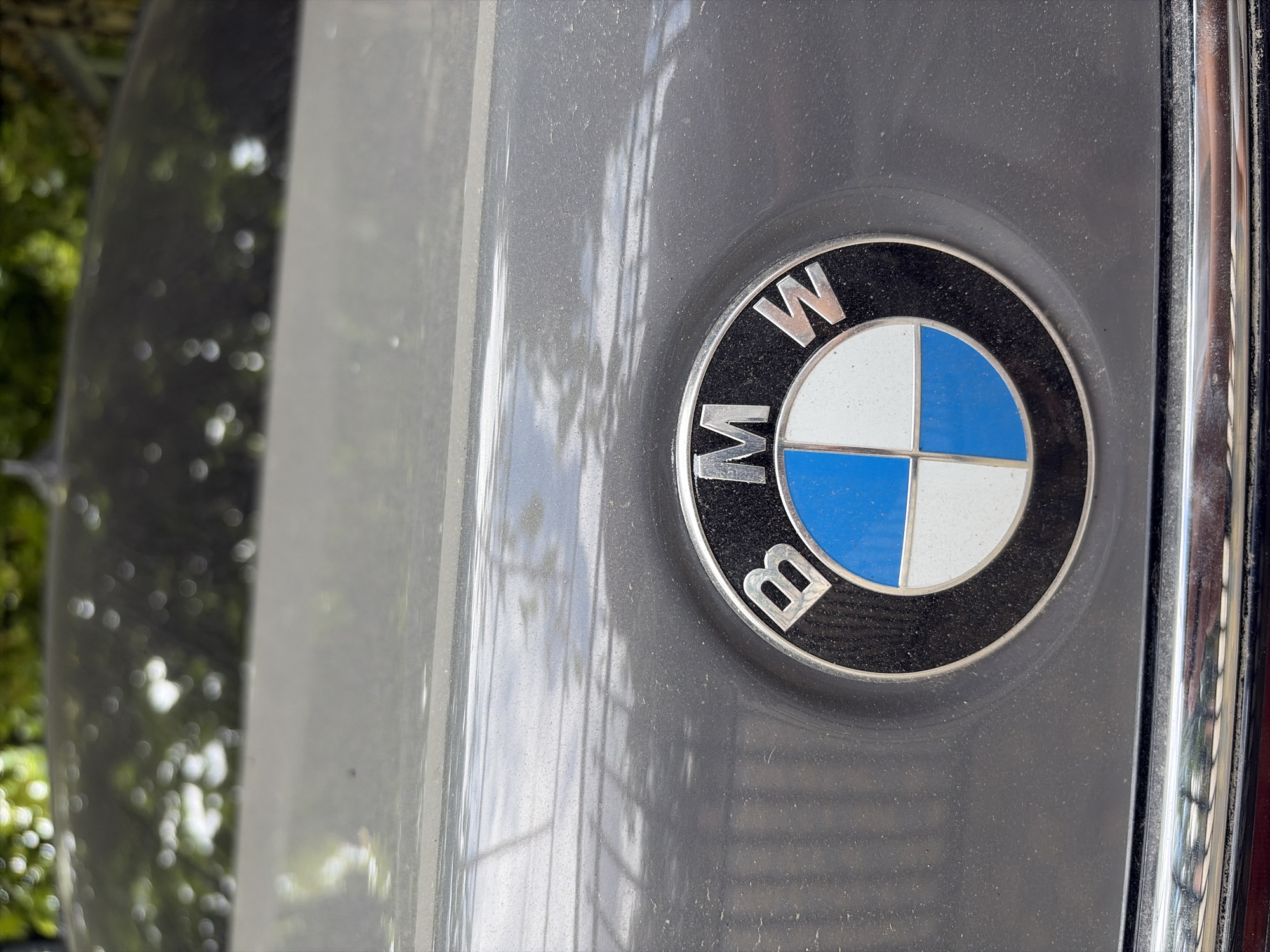 The image shows a close-up of a dusty grey BMW emblem on the trunk of a car. The emblem is circular with a black outer ring, the letters "BMW" in silver at the top, and a blue and white checkered pattern in the center, representing the Bavarian flag. The car's paint is slightly dirty, with visible dust particles. Reflections of greenery and sky are visible on the car's surface, suggesting it is outdoors. The chrome trim below the emblem is also visible and appears slightly dusty. There are no people or other significant objects in the frame. The location is stated to be Oxentea, Moldova.