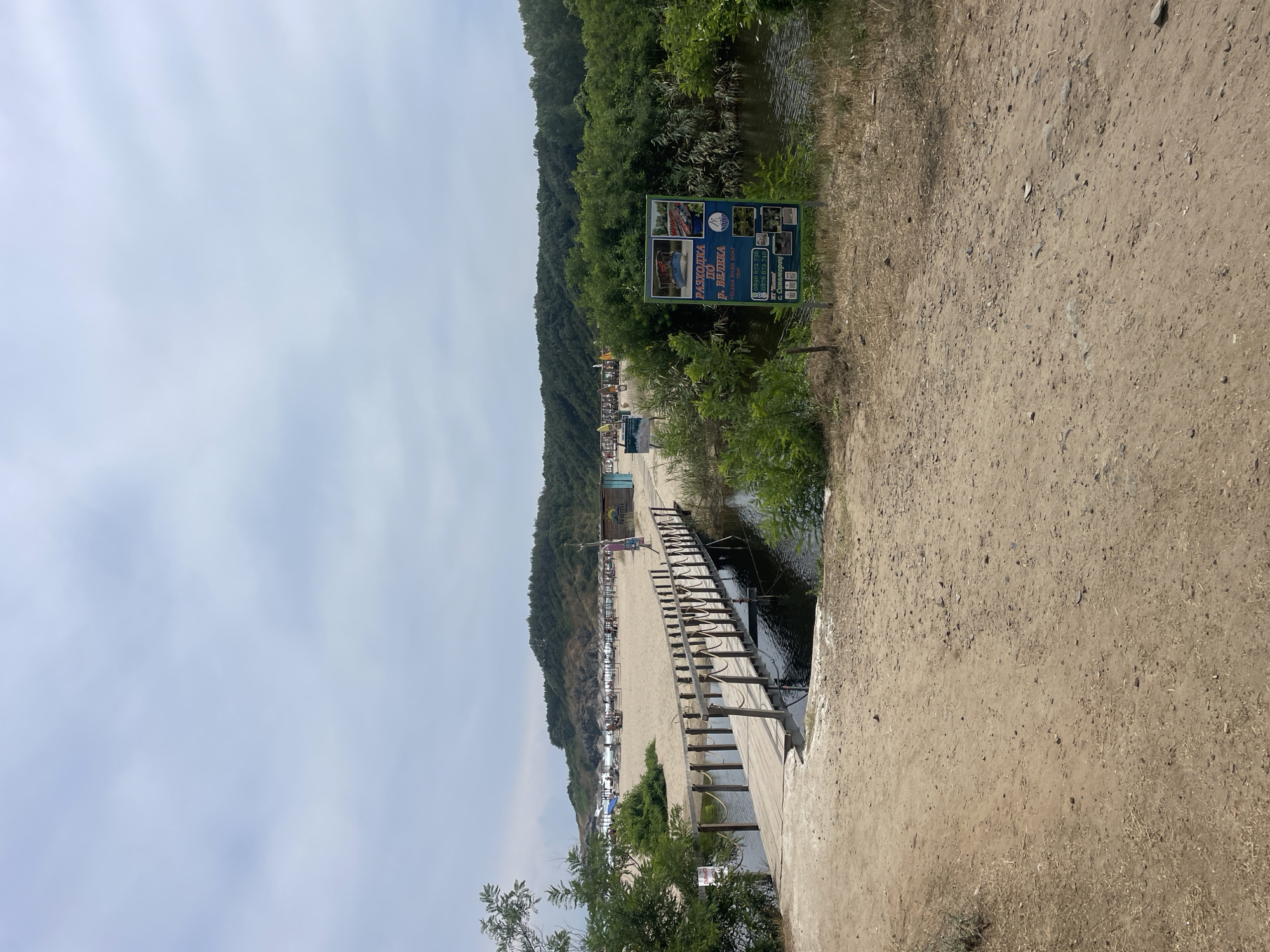 The image shows a wooden bridge crossing a small body of water that leads to a sandy beach. On the beach, there are several rows of beach umbrellas and a few small buildings. A signpost with pictures of boats and text in Cyrillic is visible on the right side of the image. The environment is natural, with trees and vegetation surrounding the area. The sky is overcast with clouds. The scene appears to be a tourist destination. Based on the information provided, the location is Rezovo, Bulgaria.