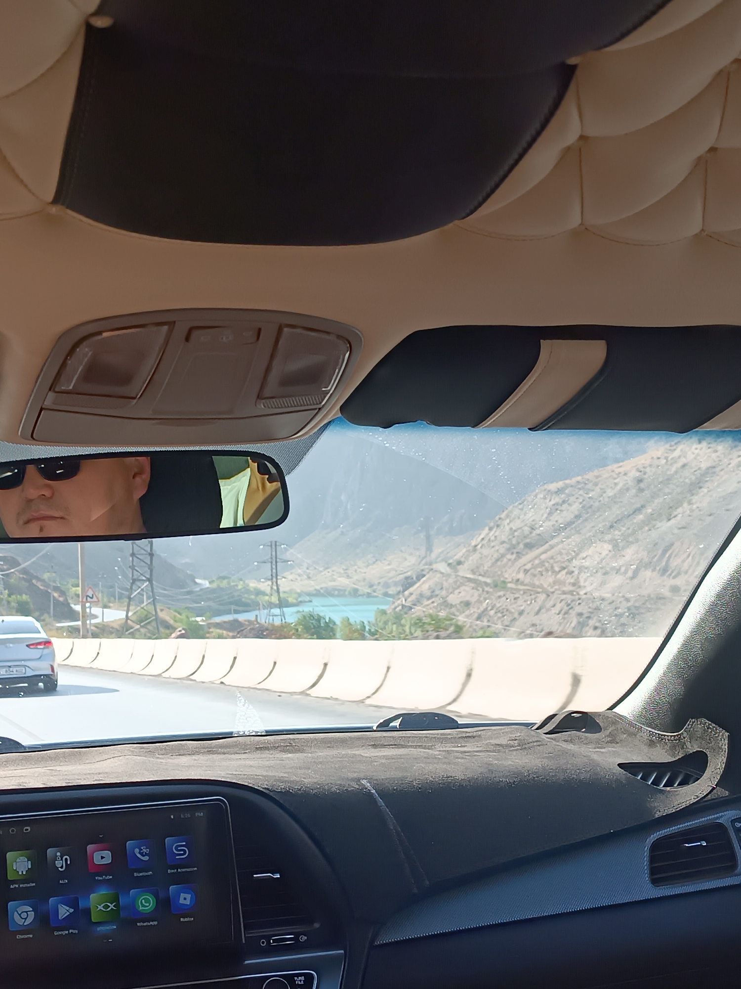 The image is taken from inside a car, looking forward and slightly upwards. The dashboard is visible in the foreground, with a dark fabric mat covering a portion of it. A car's infotainment system is displayed on a screen in the lower left of the dashboard, showing various app icons.
In the center of the image, a rearview mirror reflects a man wearing sunglasses and a goatee. Behind him, through the windshield, a scenic view of a mountainous landscape and a body of water is visible. The road ahead appears to be a highway with other vehicles present. Power lines and poles are also visible in the distance.
The overall impression is of a road trip through a mountainous region, possibly in or near Kara-Köl, Kyrgyzstan, given the context provided. The interior of the car features a padded ceiling with beige and black sections.