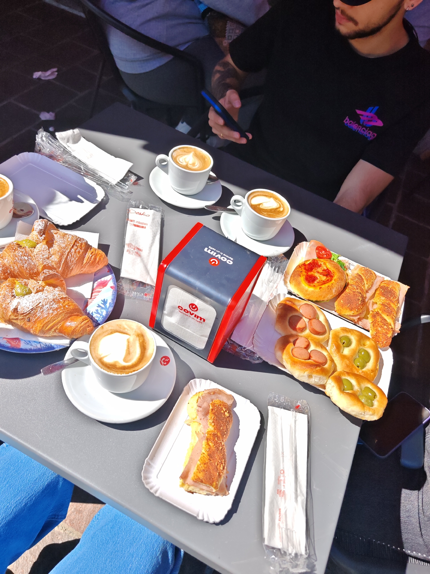 Captured from an overhead perspective, the scene portrays a gray table laden with an assortment of breakfast items, likely at a cafe in Unknown, Unknown. Dominating the view are several pastries: croissants on a blue-patterned plate, filled rolls with sausage or olives on a paper tray, and a single slice of what appears to be savory pie on another paper plate. Beverages are represented by three cups of cappuccino, each presented on a white saucer.
Additional objects on the table include a red and gray napkin dispenser labeled "COVIM Genova in un caffè," packets of sugar, wrapped straws, and paper napkins. Two individuals are partially visible, one wearing a black shirt with "balenciaga" on it and holding a cell phone, and another wearing blue pants. The table appears to be outdoors, considering the strong sunlight creating sharp shadows and the brick flooring visible in the lower-left corner.