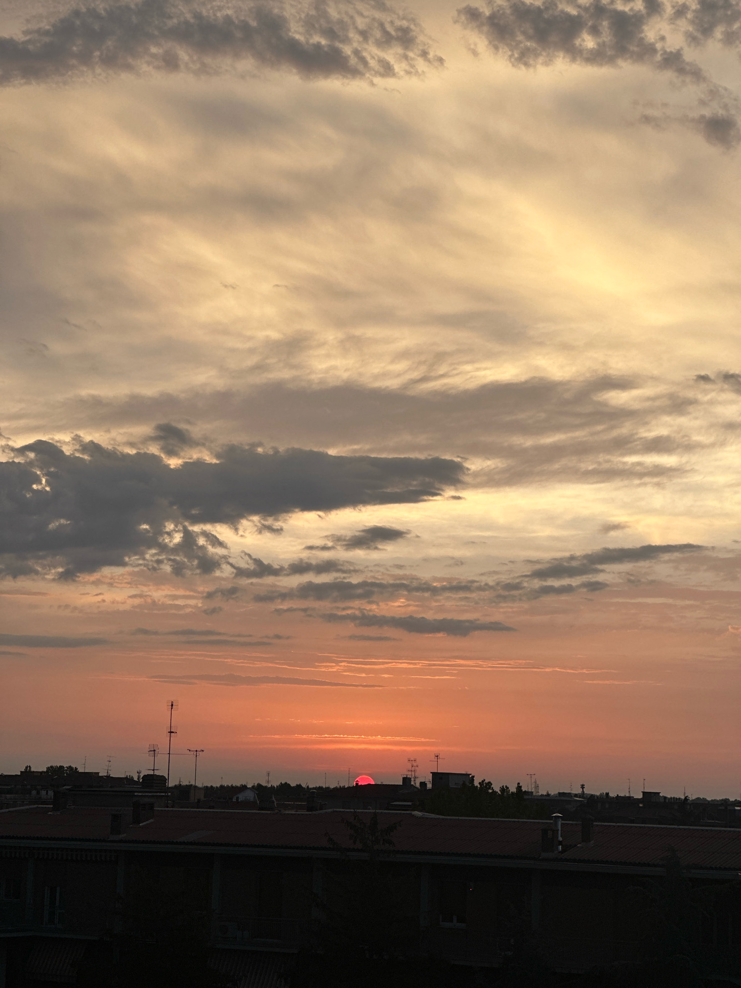 The image captures a sunset over the city of Parma, Italy. The sky is filled with dramatic clouds, painted in hues of orange, yellow, and gray. The sun, a vibrant red orb, is low on the horizon, partially obscured by clouds and the silhouettes of buildings. The foreground is dominated by the dark rooftops of urban structures, some with visible chimneys and antennas. The overall impression is one of a picturesque, yet somewhat moody, evening sky.