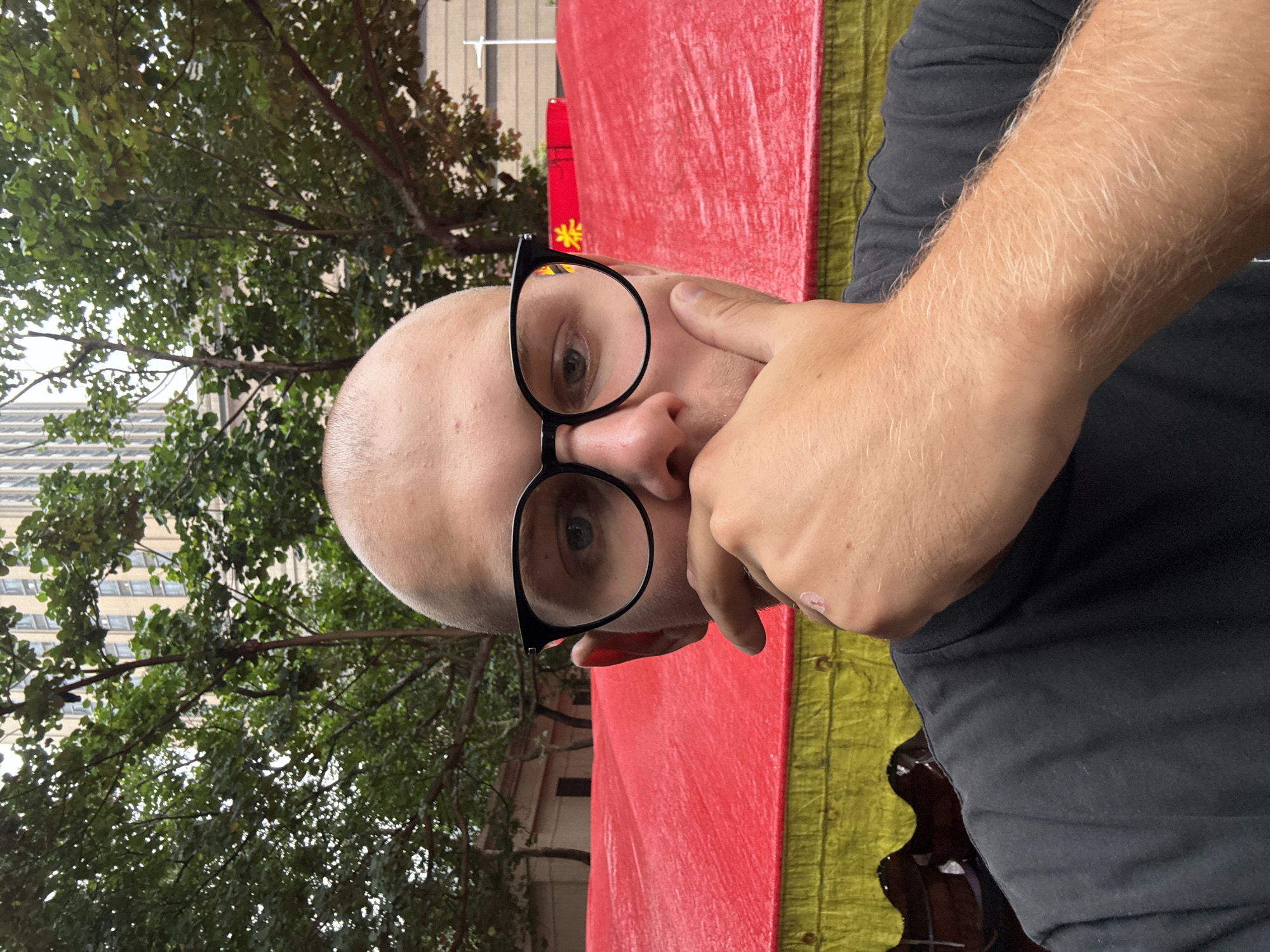A close-up, eye-level, outdoor, selfie-style shot shows a person with a shaved head wearing black-rimmed glasses and a black t-shirt. Their left hand is raised to their chin, obscuring their mouth and nose. The person's left eye is visible through the glasses, and their gaze is directed towards the camera. In the background, a large red and yellow tarp is visible, along with green trees and a tall, light-colored building. The overall impression is that the photo was taken on a street or public area, possibly during an event given the tarp. The presence of a tall building and trees suggests an urban environment.