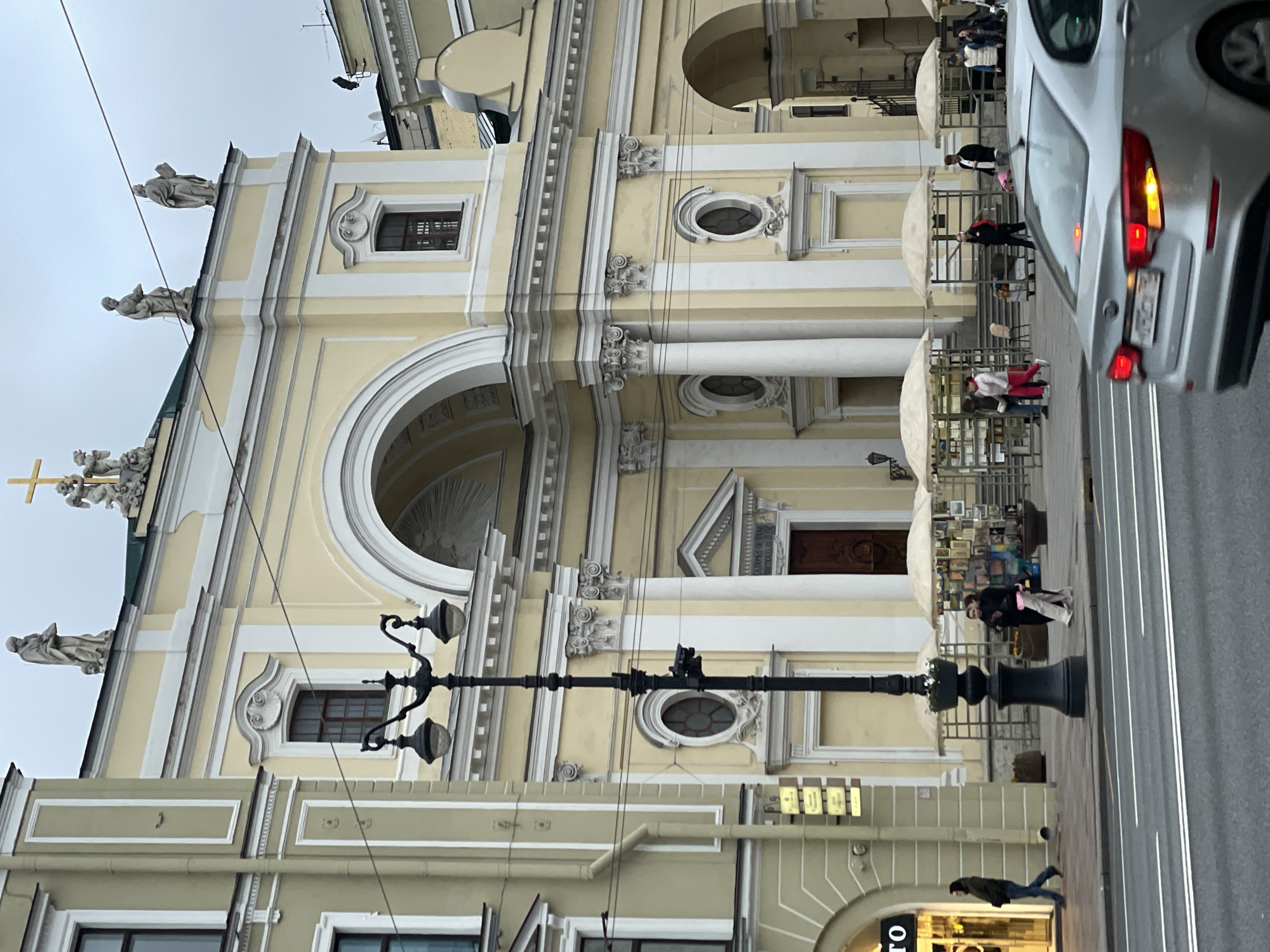 The image depicts the Church of Saint Catherine on Nevsky Prospekt in Saint Petersburg, Russia. The architecture showcases a yellow and white facade with statues on the roof, arched windows, and a large arched entrance. The street features a black lamppost, pedestrian traffic, and a car. The scene is set during the day under a cloudy sky.