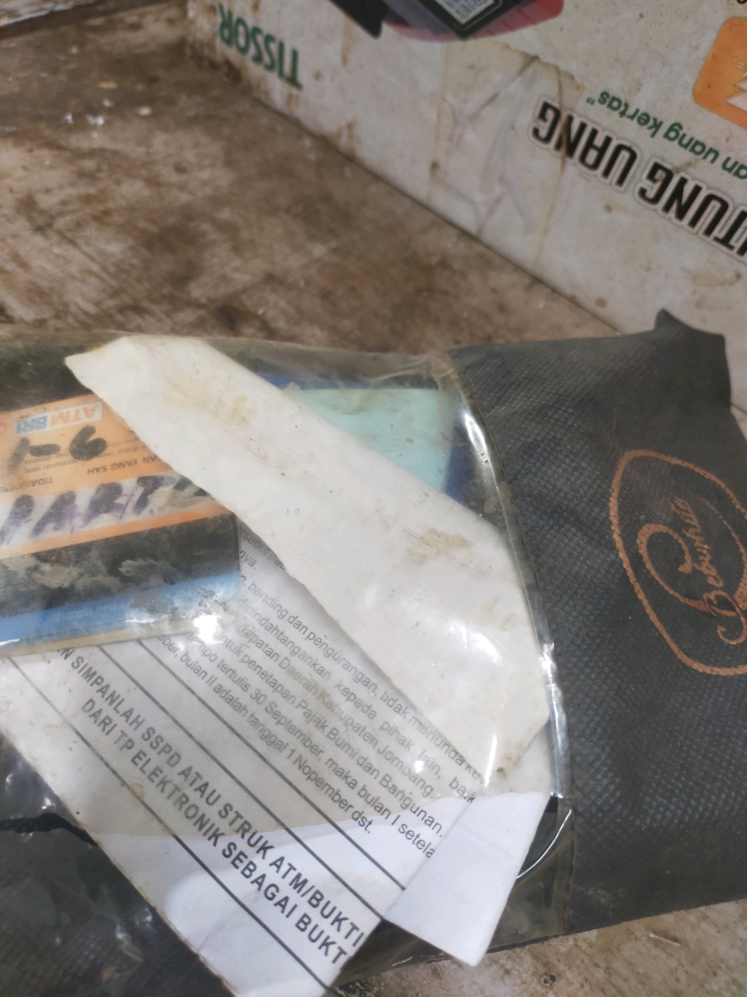 The image shows a close-up of a plastic-wrapped item containing several documents and what appears to be a black pouch with a circular emblem. The documents are mostly white with black text. Some visible text includes "TISSOR", "ATM BRI", "YANG SAH", "TIDAK SAH", "PART", "N SIMPANLAH SSPD ATAU STRUK ATM/BUKTI DARI TP ELEKTRONIK SEBAGAI BUKTI", and text in Indonesian that mentions "Pajak Bumi dan Bangunan" and dates. The black pouch has a textured surface and an oval-shaped emblem with intricate lettering. The overall scene appears to be indoors, with a dusty, textured surface visible in the background. There are no people or animals present. The image is a static shot, providing no information about actions or interactions. The location is not explicitly mentioned in the image itself.