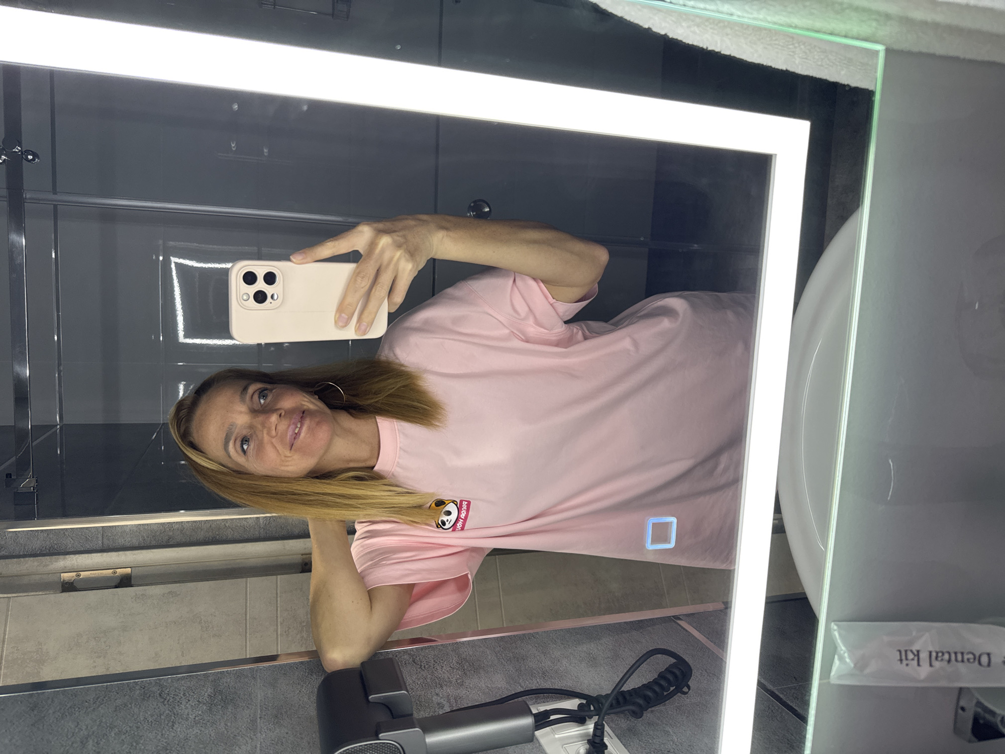 The image is a selfie taken in a bathroom mirror. A woman with blonde hair is holding up a cell phone, a modern iPhone with a beige case. She is wearing a pink t-shirt with a cartoon graphic. The bathroom features gray tiles and modern fixtures, including a sleek gray hair dryer. A white sink is visible, along with a dental kit. Based on the details visible in the image, it is impossible to determine the exact location within Tashkent, Uzbekistan.