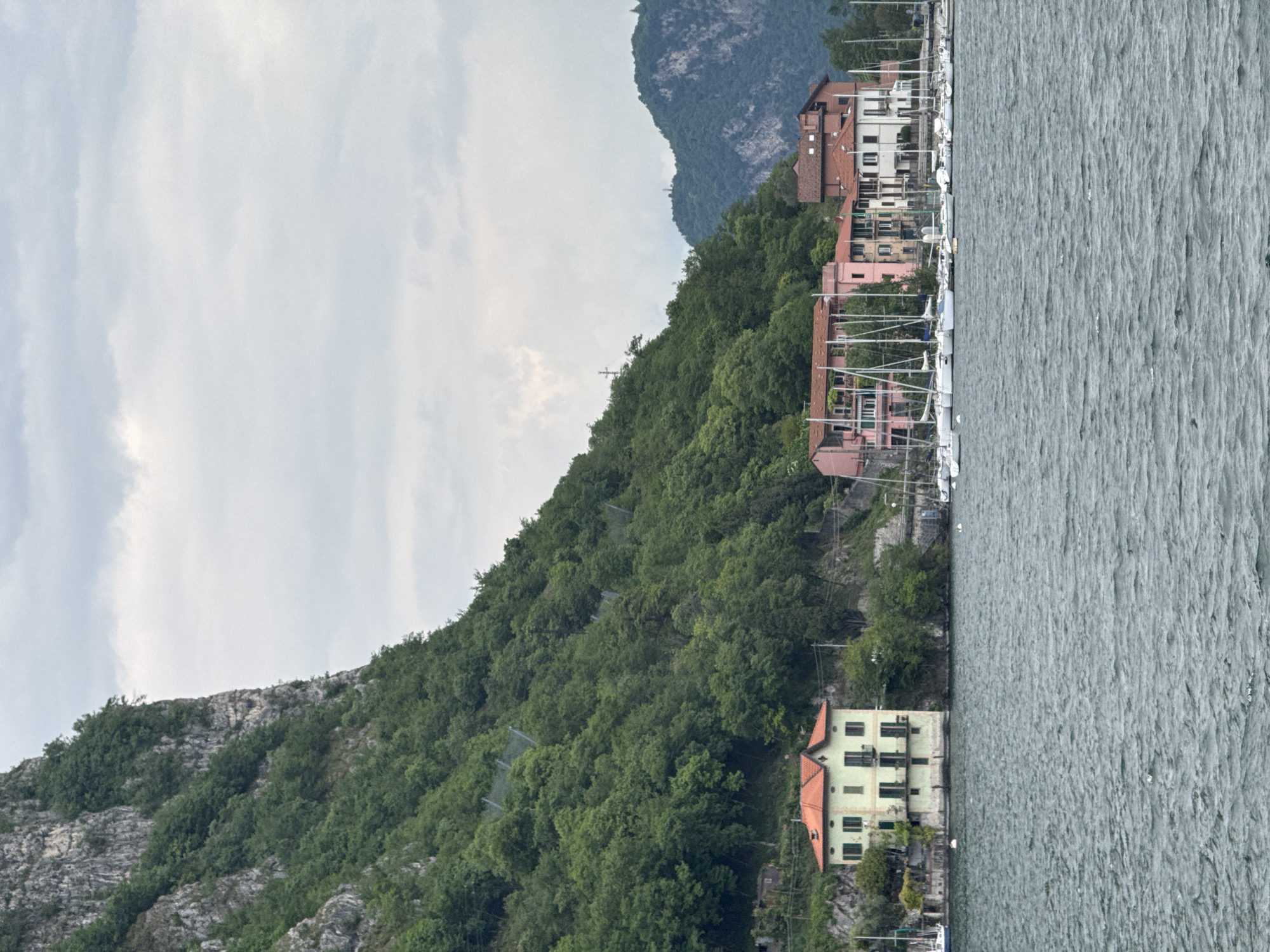 The image, when viewed upright, presents a picturesque landscape of Valmadrera, Italy, showcasing a lakeside scene under an overcast sky. In the immediate foreground, the surface of a large body of water, likely Lake Como, is visible, characterized by its choppy, grey-green ripples.
Along the lake's edge, a charming settlement unfolds with a line of multi-story buildings. These structures feature traditional terracotta roofs and facades in varying colors, including pink, white, and a light green or yellow hue. In front of these buildings, numerous sailboats are moored, indicating a marina or small port.
Behind the lakeside settlement, a steep, heavily forested hillside rises sharply, covered densely with green trees. Faint lines of what appear to be rockfall netting or erosion control measures are visible on parts of this slope, and a small cross can be discerned towards its upper section. Further in the background, a larger, rugged mountain range, partially covered in vegetation and exposed rock faces, dominates the horizon.
The sky is uniformly overcast with soft, diffused clouds, suggesting a muted, daylight ambiance without direct sunlight. There is no explicit activity taking place, and no visible text can be identified within the image.