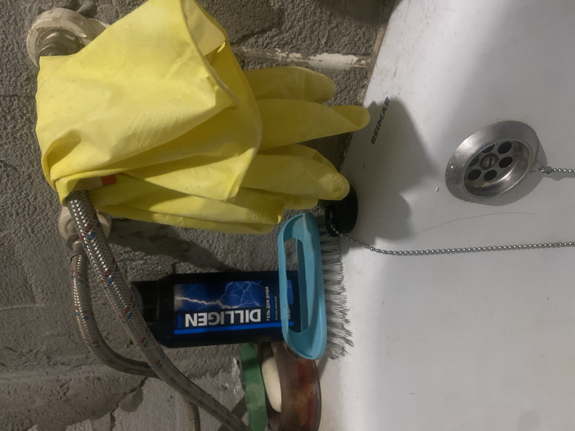 A close-up, waist-level shot captures a bathroom sink area with various cleaning and personal hygiene items. A white ceramic sink basin dominates the foreground, with a chrome drain cover featuring multiple circular holes in the center. A thin, silver beaded chain hangs from the drain cover, disappearing downwards. To the left of the drain, a blue-handled scrubbing brush with white bristles rests on the edge of the sink. Behind the brush, a clear plastic soap dish holds a bar of white soap and a green sponge. A black bottle with a blue label that reads "DILLIGEN" in white text is positioned behind the soap dish. The bottle has an image of blue lightning bolts. Above the sink, a flexible metal hose, likely for a faucet, is visible, supporting a folded yellow cloth or pair of gloves. The background consists of a textured, gray concrete wall. The scene appears to be indoors, in a bathroom.