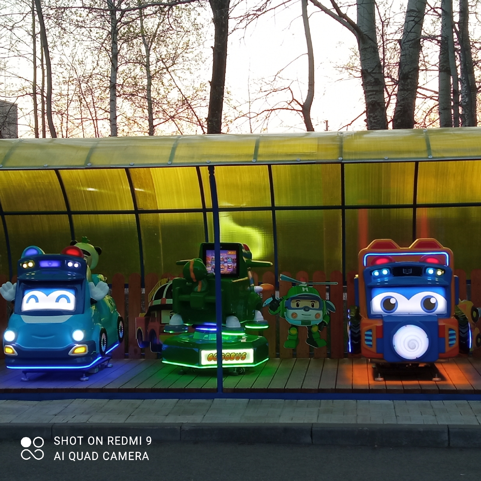 The image shows three coin-operated kiddie rides under a transparent yellow roof in Chaikovsky, Russia. The rides are on a wooden platform bordered by a low wooden fence. From left to right, the rides are: a blue car with cartoon eyes and a white mouth, a green helicopter with a screen displaying images, and an orange and blue machine with a large circular light. In the background are trees with bare branches against a sky transitioning from yellow to pale blue. The foreground shows a gray concrete curb. The photo includes a watermark in the lower left corner.