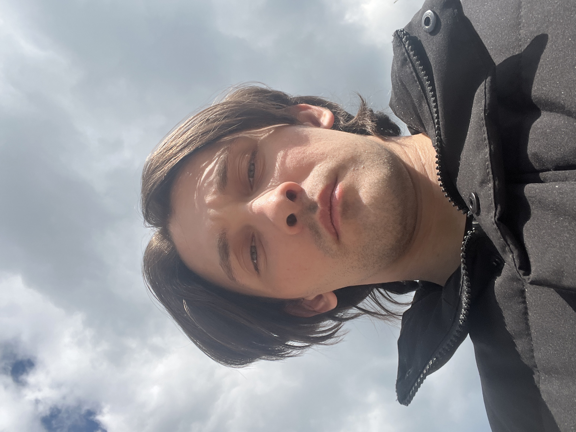 An extreme low-angle, medium-close-up selfie shows a man's face against a cloudy sky. The man has medium-length, dark brown hair, parted in the middle and falling to his ears. He has fair skin, prominent cheekbones, and a slight stubble. His eyes are open and looking forward, and his lips are closed. He is wearing a black puffer jacket with a high collar that is zipped up. The background is dominated by a pale blue sky with scattered white clouds. The image appears to have been taken outdoors in the city of Orekhovka, Russia.