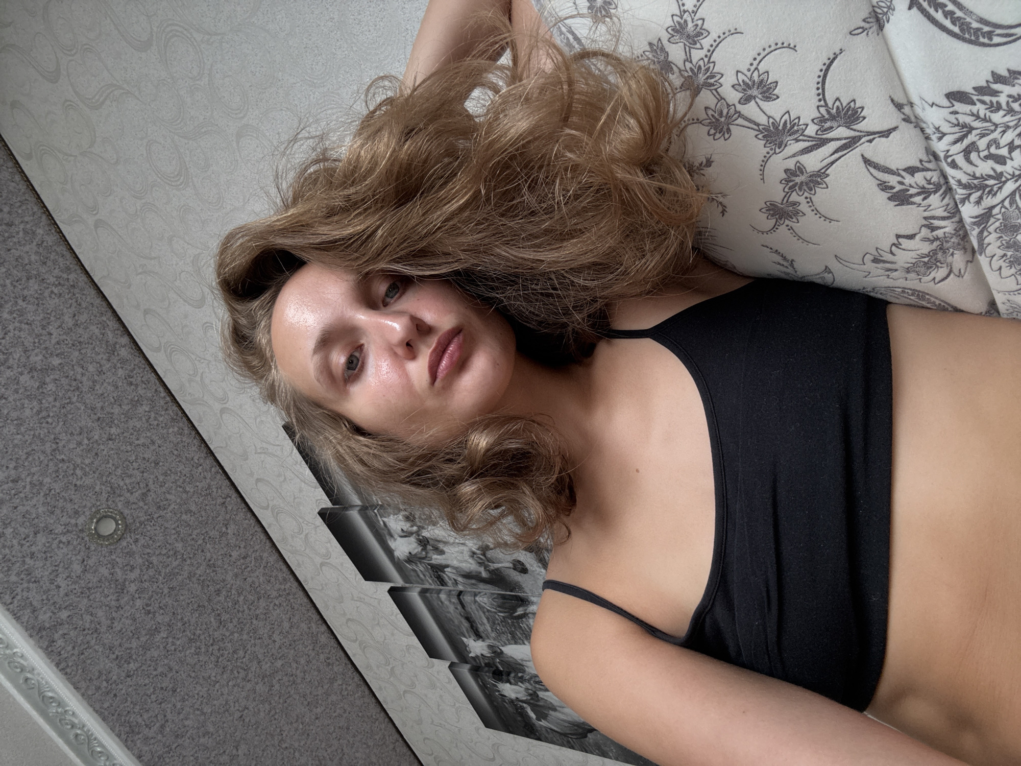 A young woman with blonde, wavy hair is lying down, looking towards the camera. She is wearing a black tank top. Her right arm is resting behind her head, partially obscuring her hair. In the background, there is a wall with a patterned wallpaper and a multi-panel artwork of horses. A textured ceiling with a light fixture is visible in the upper left corner. The scene appears to be indoors, possibly in a residential setting. There is no information in the image to determine the specific location within the city of Kuyeda, Russia.