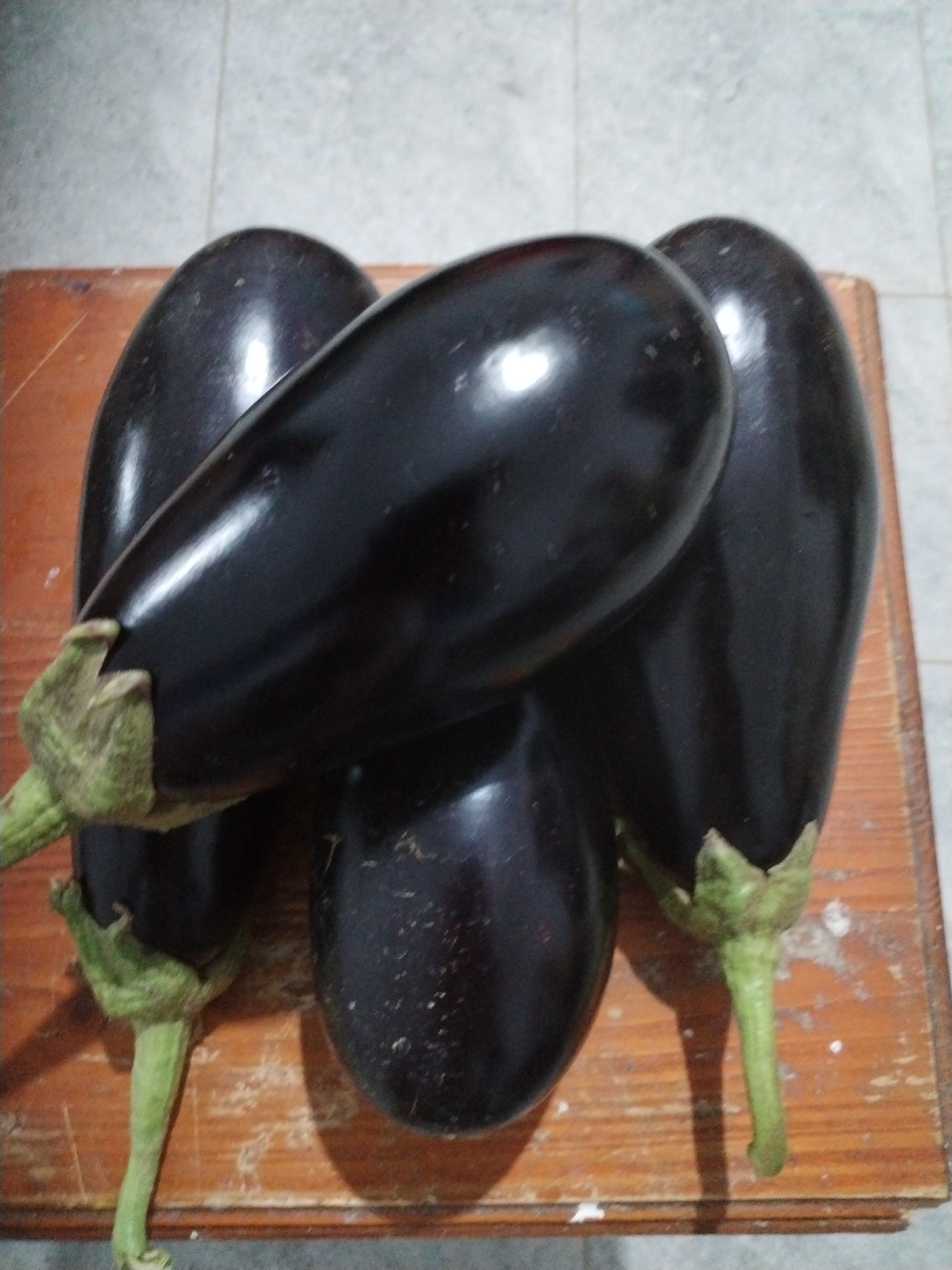 The image displays four dark purple eggplants arranged on a worn wooden surface. The eggplants are of varying sizes and shapes, with some exhibiting a glossy sheen. One eggplant is prominently positioned in the foreground, dominating the central area of the frame. The wooden surface appears to be a table or countertop with visible grain and some lighter-colored marks. In the background, a tiled floor is partially visible, with the tiles appearing to be a light gray color. The lighting suggests an indoor environment, possibly a kitchen or pantry. There are no people or animals present in the image, and no specific actions or interactions are depicted. The scene is static and focuses solely on the arrangement of the eggplants. The image was taken in Moara Domnească, Romania.