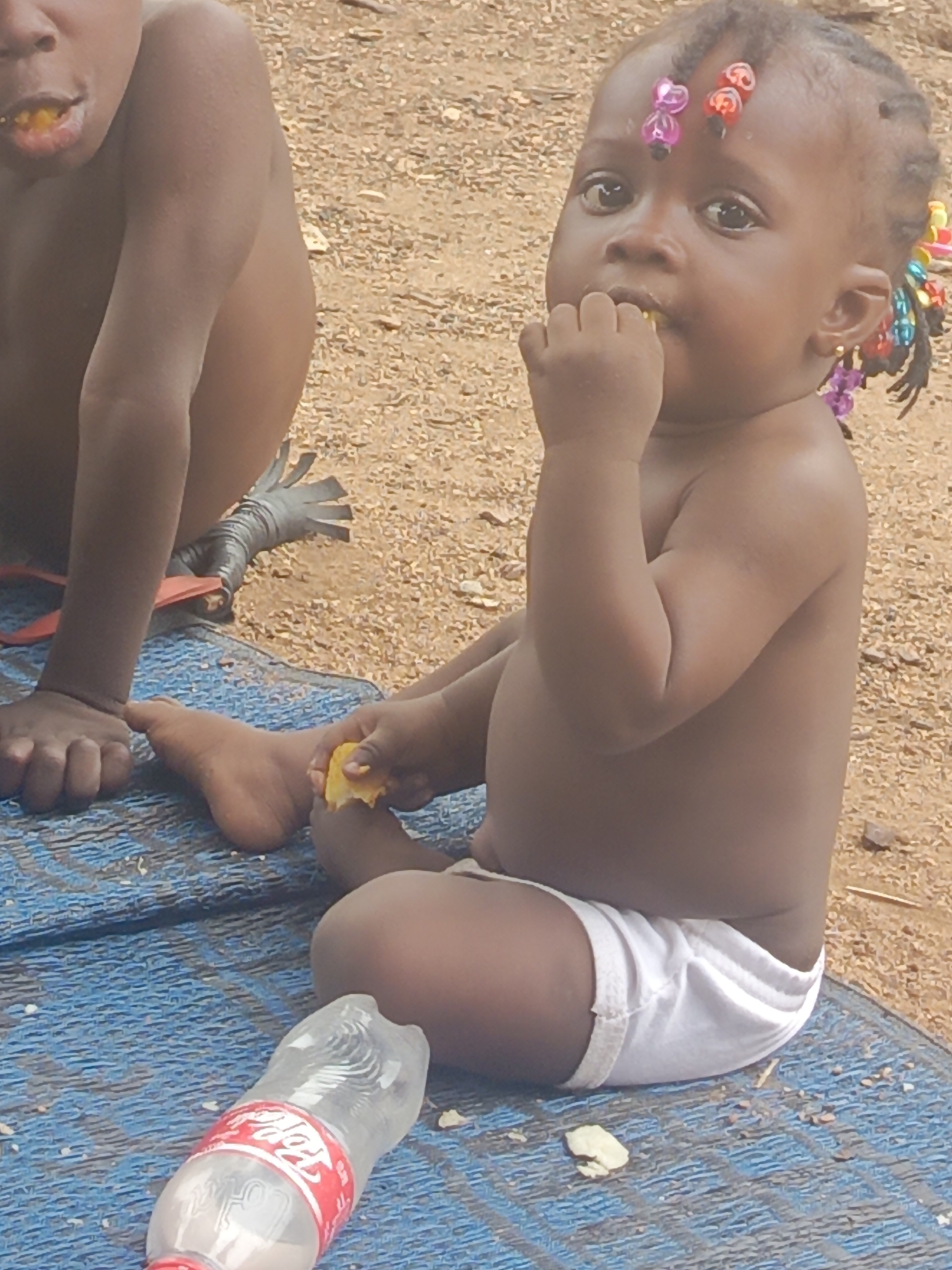 Two dark-skinned children are seated on a blue patterned mat placed on sandy ground. A baby girl with hair adorned with colorful plastic flowers and beads is eating, holding food in both hands. Next to her is another child, partially visible, also eating. A clear plastic bottle with a red label, likely for a Coca-Cola product, lies on the mat. The location is Akwanga, Nigeria.