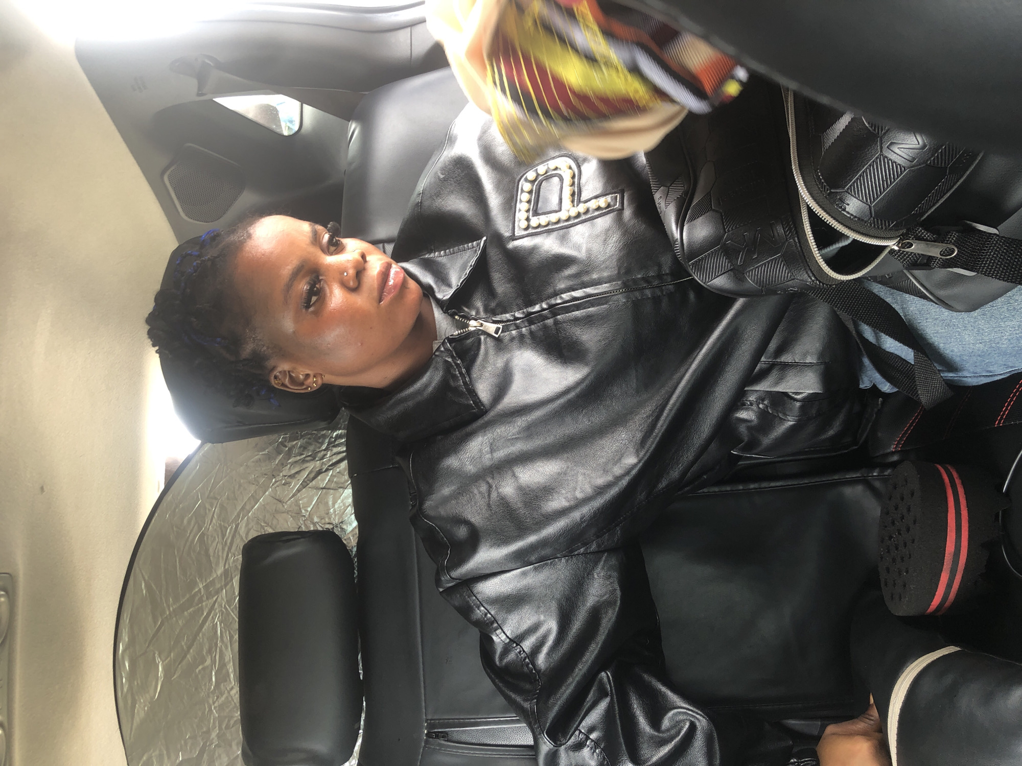 A young Black woman with braided hair, adorned with blue extensions, sits in the backseat of a car. She is wearing a black leather jacket with a pearl-encrusted letter "P" on the left breast, and a grey t-shirt underneath. Her head is tilted down, and she appears to be looking at something in her lap. A black backpack with a textured pattern and a zipper partially open rests beside her. On the floor of the car, in front of her, is a black, circular hair sponge with red stripes. The car's interior is visible, including black leather seats and the headrest of the seat in front of her, which is partially obscured by a silver sunshade. The scene takes place inside a vehicle, likely in Ikoyi, Nigeria, suggesting an urban setting.
