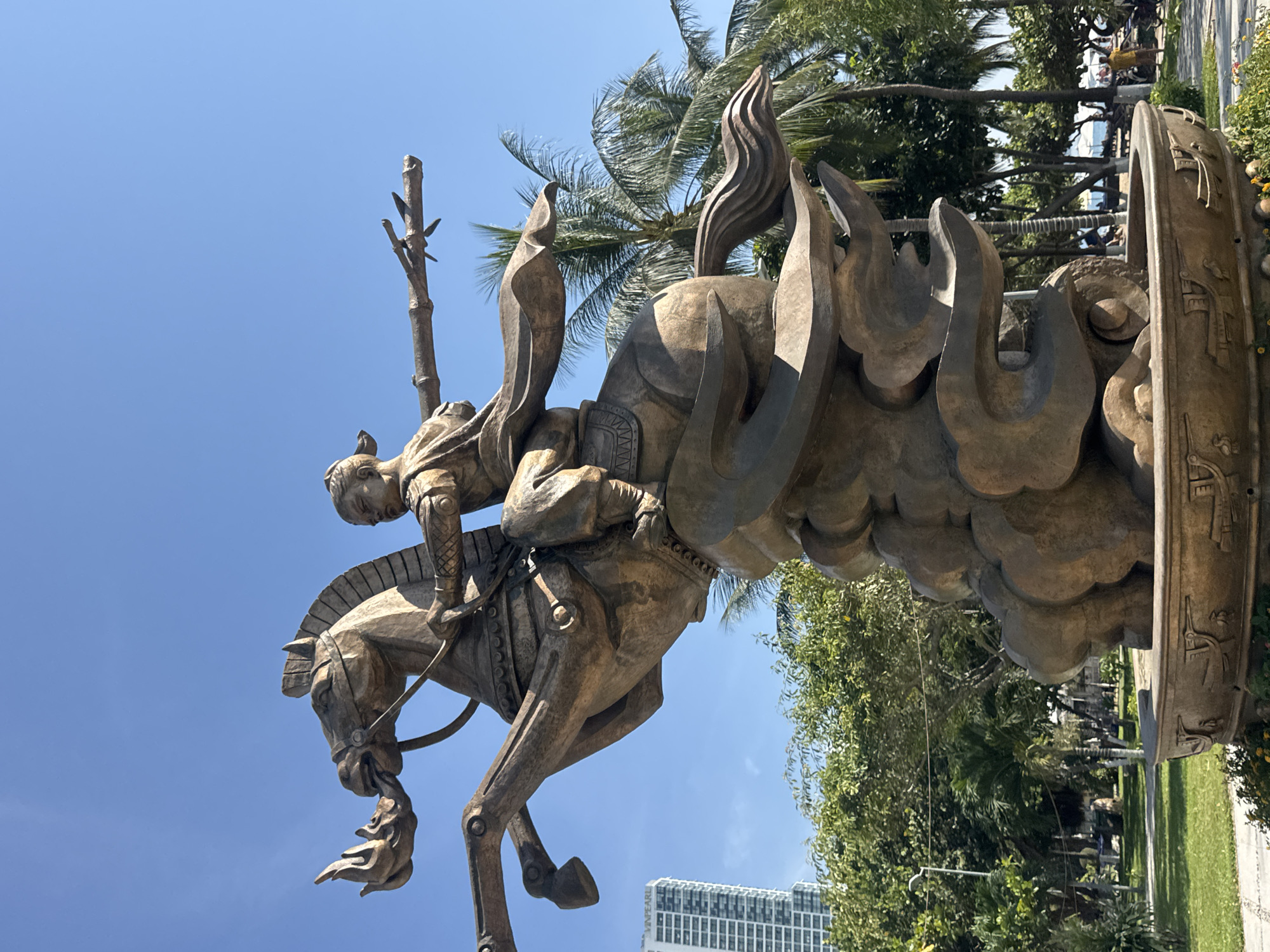 A bronze statue depicts a warrior on horseback, with the horse rearing up on a cloud-like base. The warrior, dressed in traditional attire with a flowing sash, holds a bamboo pole outstretched to the right. The horse has a fiery mane. In the background, palm trees and lush greenery are visible, along with a tall, modern building labeled "VINPEARL" and a clear blue sky. The statue appears to be located outdoors in a park-like setting, possibly a public square or promenade, with a pathway and hints of a beach in the distance. This is the Tran Hung Dao statue located in Nha Trang Ward, Vietnam.