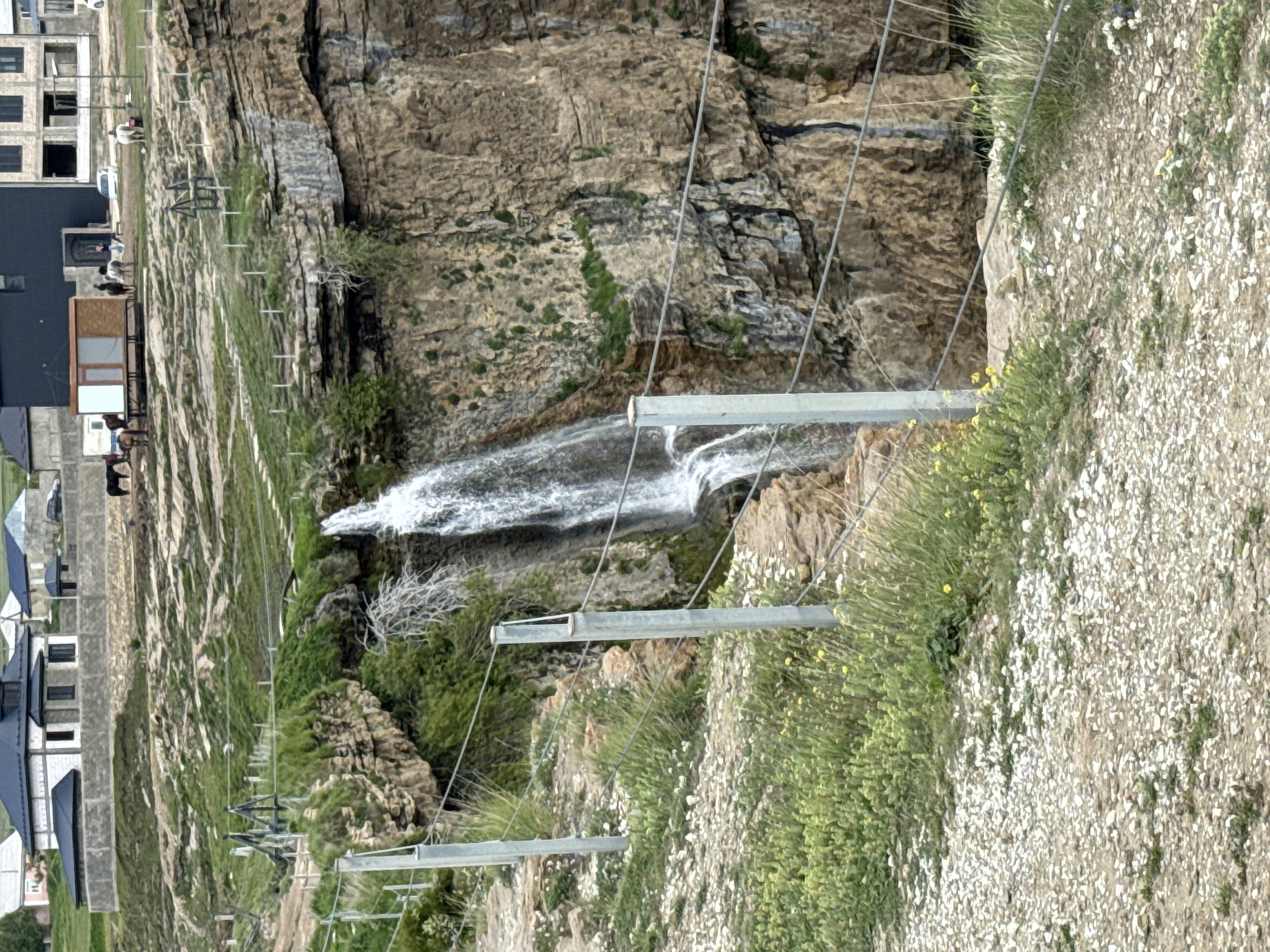 This image captures a scenic landscape featuring a waterfall cascading down a rugged, rocky cliff. The scene is set in a mountainous or plateau region, with human structures visible on the flat terrain above the cliff.
At the top of the cliff, several buildings are present, some appearing to be under construction with exposed concrete and stone-like facades, while others have dark, finished roofs. Nearby, a group of horses is visible, standing on the grassy ground, with a small wooden structure or shelter positioned among them. A verdant, grassy expanse covers the plateau's edge.
The focal point is the powerful waterfall, which plunges over the steep, stratified rock face. The cliff itself displays varied textures and shades of brown and gray rock, with scattered patches of green vegetation and small trees growing from its crevices.
In the foreground, a series of robust metal posts with horizontal bars and multiple taut cables stretch across the scene. These elements suggest a protective barrier, a viewing platform, or perhaps part of a specialized infrastructure, like a suspension bridge or a recreational installation, providing a clear vantage point towards the waterfall and cliff. The immediate ground cover consists of sparse green grass dotted with numerous small white and yellow wildflowers, leading to a patch of gravelly earth.
The lighting indicates daytime, with soft, diffused light, possibly from an overcast sky. There are no visible people or discernible activities other than the horses grazing. No visible text is present in the image. The location is contextually identified as Khunzakh, Russia.
