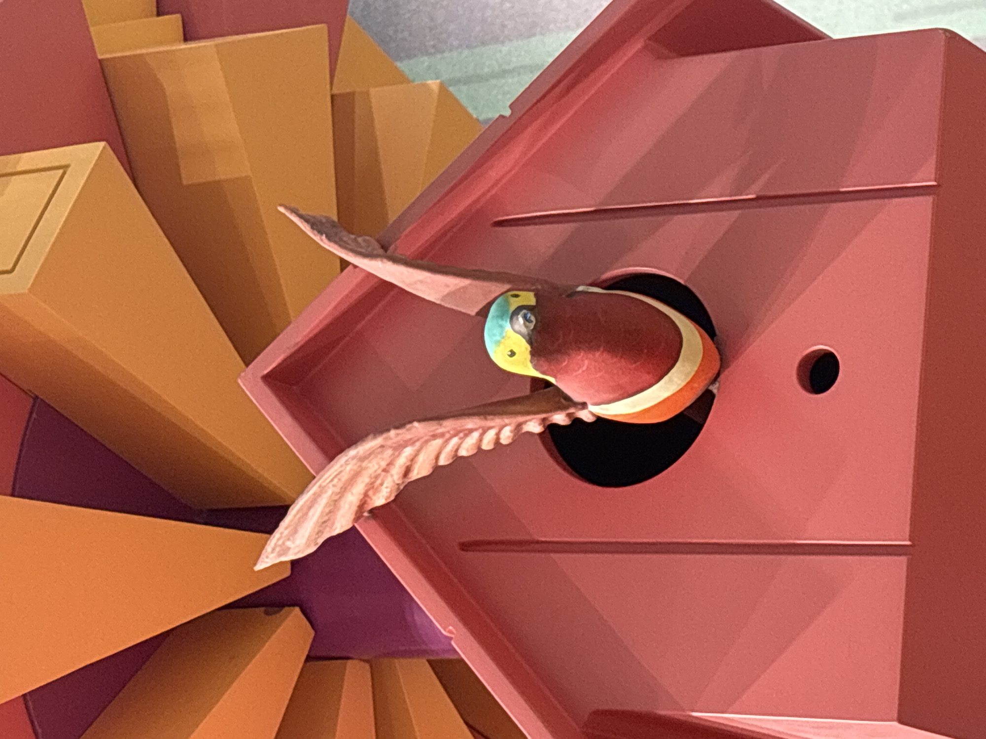 A close-up shot focuses on a red birdhouse, with a colorful toy bird emerging from the opening. The bird has a red body with white and orange stripes around its neck, and a blue, yellow, and green head. Its wings, which are detailed with feather textures, are spread wide. The birdhouse itself is a vibrant red, with a circular opening and a smaller hole below. Behind the birdhouse, a backdrop of orange and purple geometric shapes creates a dynamic, layered effect. The scene appears to be indoors, possibly a display or part of a playful installation. There are no people or animals present in the image.
