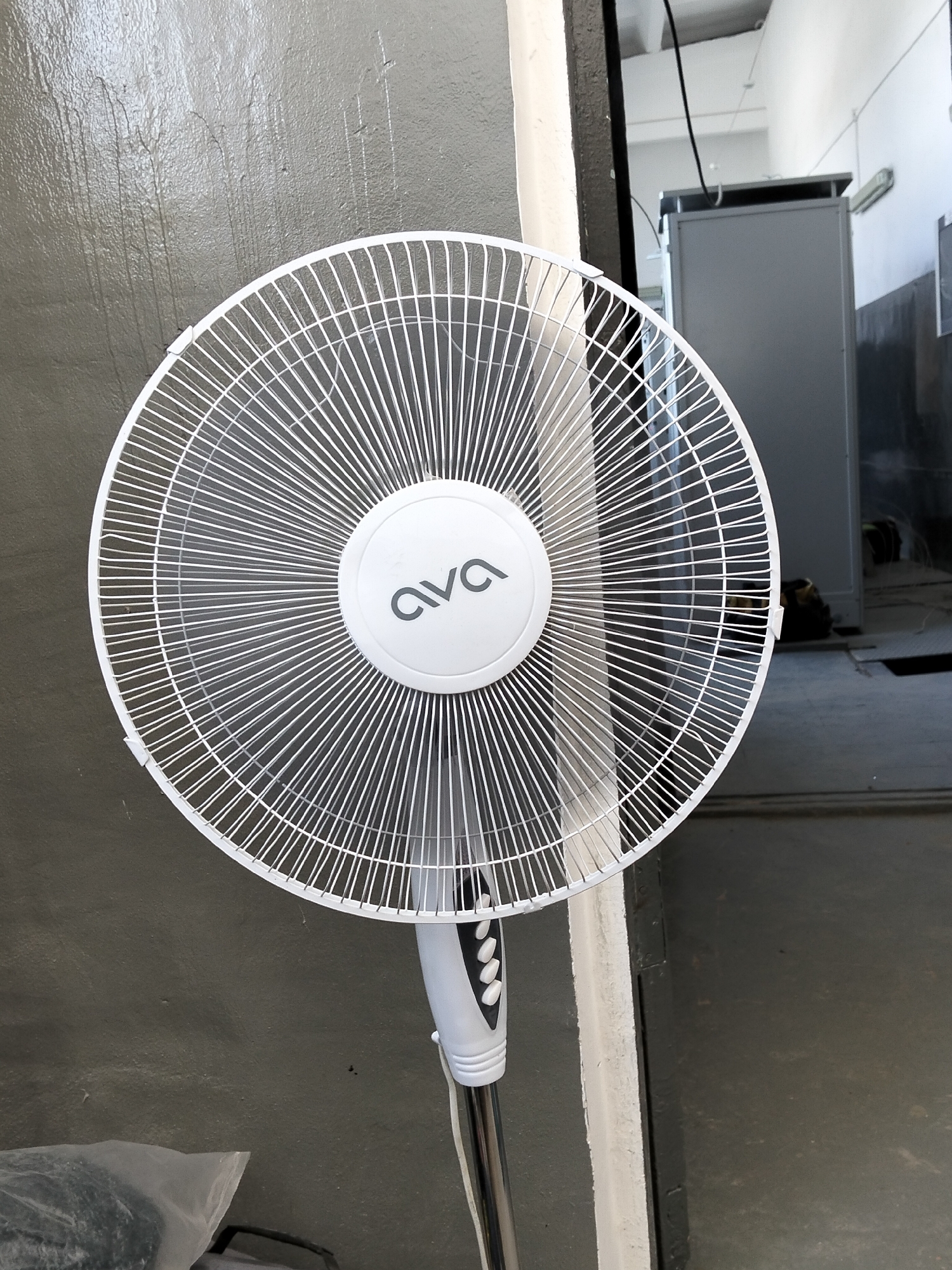 An indoor shot shows a white pedestal fan with the brand name "ava" visible on its central hub. The fan is positioned in front of a grey, textured wall. To the right of the fan, a doorway leads to an area with industrial-looking equipment, including a grey metal cabinet and some electrical wiring. The floor in the background appears to be concrete. In the lower left corner, a clear plastic bag containing dark items is visible. The scene suggests an interior space, possibly a workshop or storage area.
