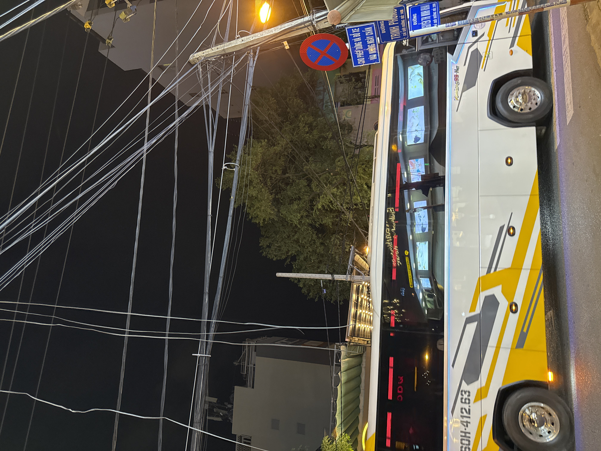 A bus with license plate "60H-412.63" is parked on a street at night in Nha Trang, Vietnam. The bus is white with yellow and gray geometric accents. The bus has several screens visible through its windows, displaying what appear to be images or videos. There are also overhead power lines and streetlights. A red and blue traffic sign is visible on the right side of the image, along with blue street signs. The street is paved, and a crosswalk is visible in the foreground. The scene appears to be in an urban area.