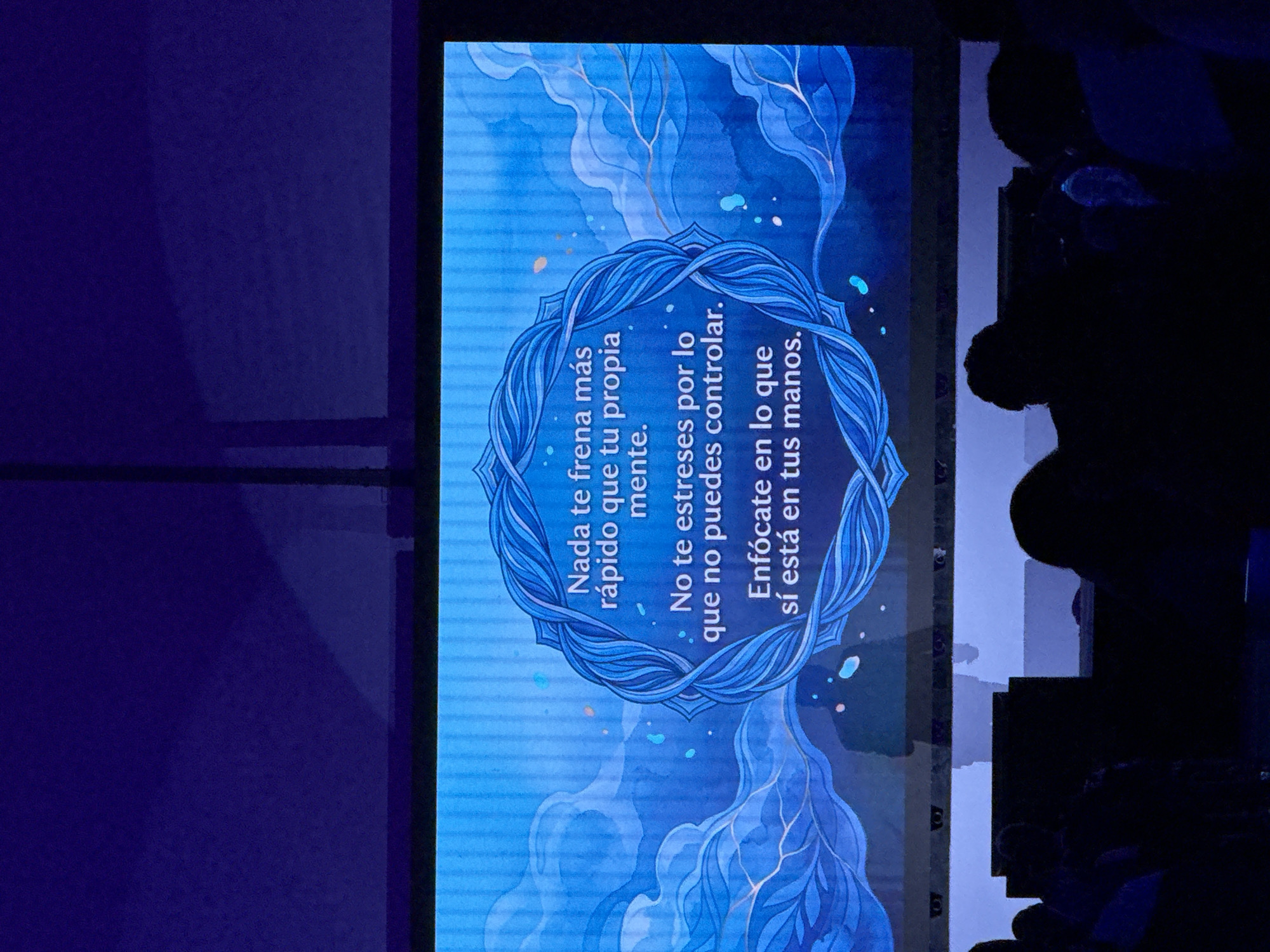 The image shows a screen displaying text in Spanish set against a blue, watercolor-like background with a stylized floral or fractal design. The text reads, "Nada te frena más rápido que tu propia mente. No te estreses por lo que no puedes controlar. Enfócate en lo que sí está en tus manos." (Nothing slows you down more than your own mind. Don't stress about what you can't control. Focus on what is in your hands). To the right of the screen, there are silhouettes of people's heads and shoulders, suggesting they are observing the screen or presentation. The overall lighting appears subdued.