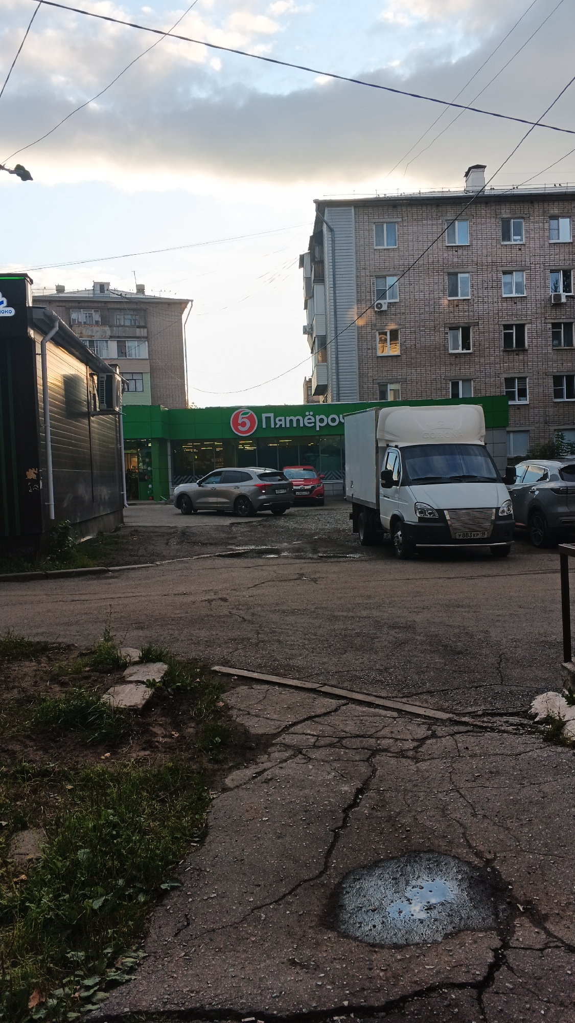 The image depicts an outdoor scene in Izhevsk, Russia, likely in a residential or commercial area. In the foreground, a cracked asphalt surface with patches of grass and a puddle reflecting the sky is visible. To the right, a white truck with a box body is parked. In the mid-ground, a silver SUV and a red car are parked in front of a bright green building with the "5" logo and "Пятёрочка" text, indicating it is a Pyaterochka grocery store. Behind the store, a multi-story brick apartment building stands. To the left of the grocery store, a dark-colored building with a reflective facade is partially visible. The sky above is partly cloudy, with the sun appearing to be setting or rising. No people are clearly identifiable in the scene. The overall impression is of a typical urban environment in Russia.
