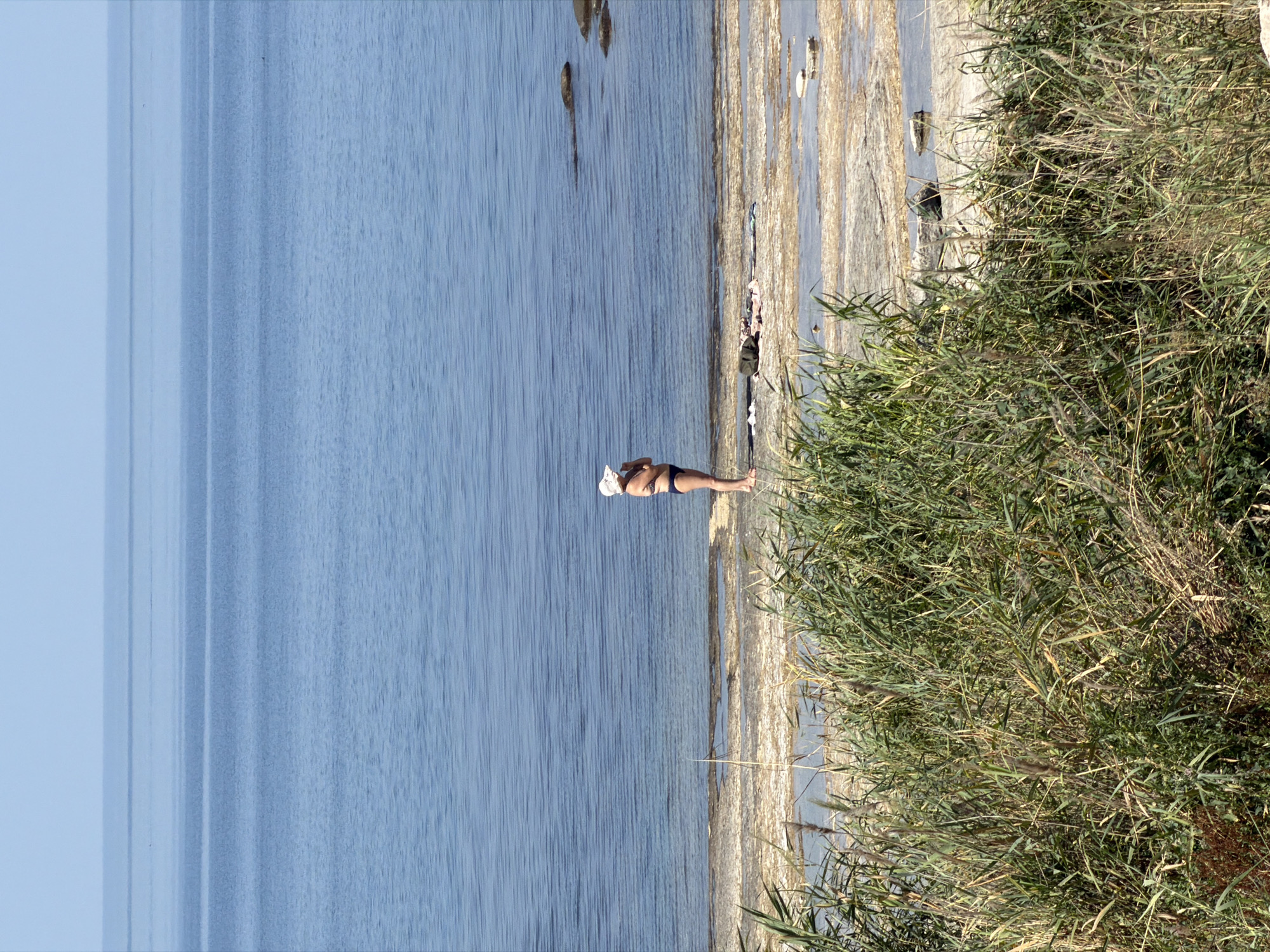 A woman wearing a white wide-brimmed hat and a dark bikini stands on a rocky, shallow shore of a large body of water. The water, a deep blue, stretches to the horizon, meeting a light blue sky with no visible clouds. The shore is a mix of light-colored sand, rocks, and shallow pools of water. Reeds and tall grasses grow in the foreground, obscuring the lower part of the image. The scene is peaceful and still, with the woman appearing to be looking out at the water. This location is likely near Aktau, Kazakhstan, a city known for its Caspian Sea coastline.