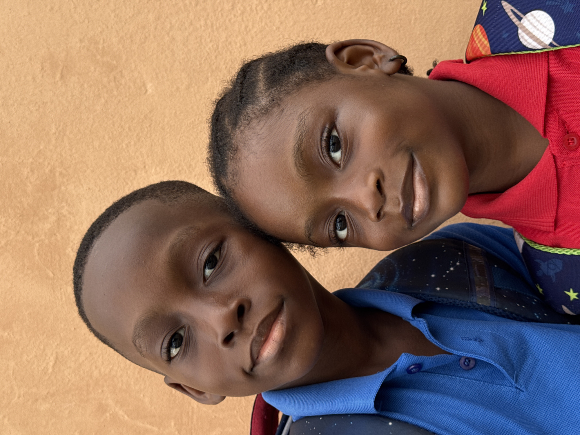 Two dark-skinned children are closely posed for a portrait in front of a plain, light-brown wall. The child in the foreground wears a blue collared shirt and a backpack with a space-themed pattern. The child behind wears a red collared shirt with a space-themed backpack. The location, Ado, Nigeria, can only be inferred, not forensically determined from the image itself.