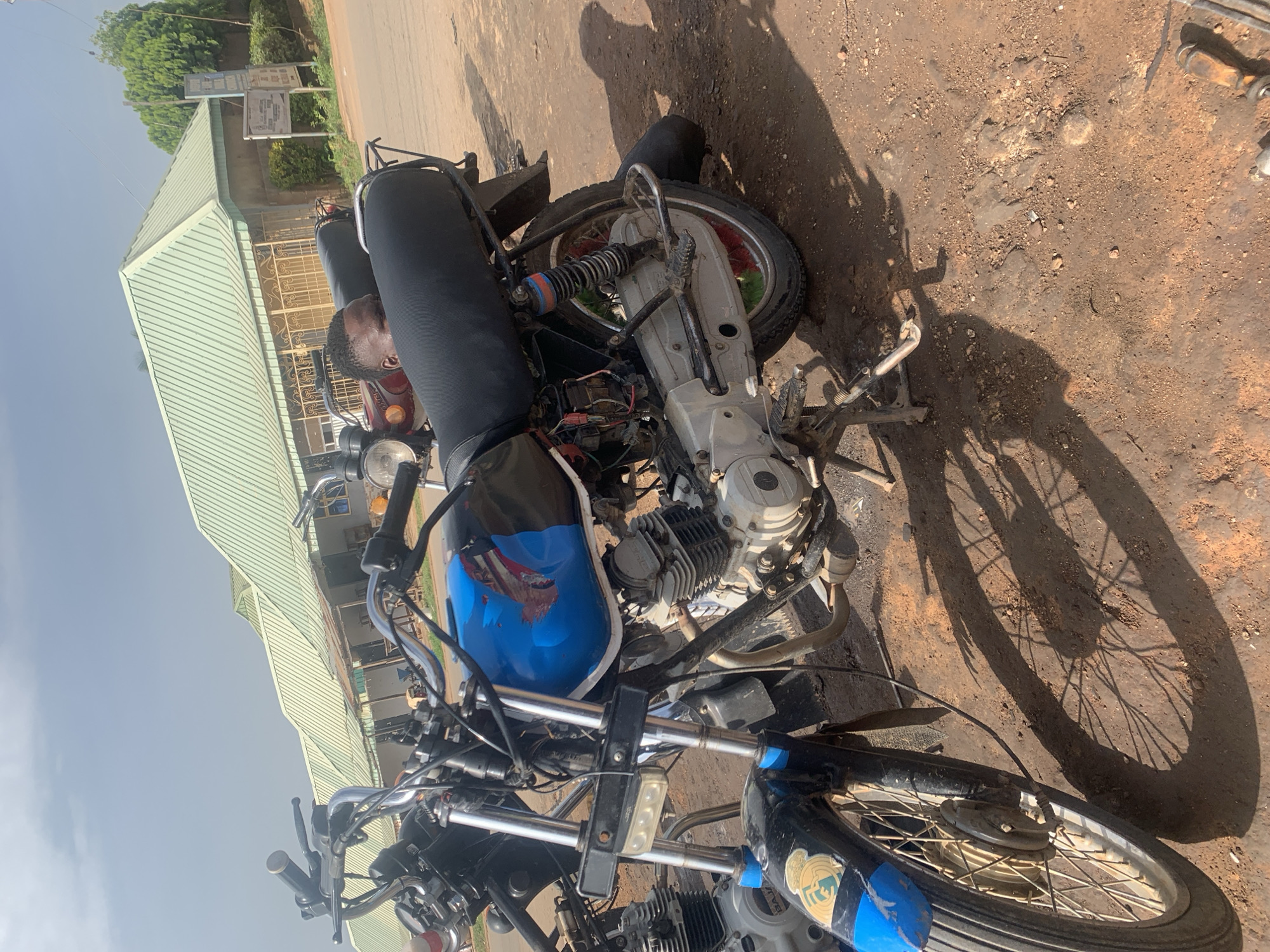 In Gura Top, Nigeria, two motorcycles are parked outdoors on a dirt road in front of a building with a green, corrugated metal roof. The motorcycle in the foreground is blue and black with a decal on its fender. A person is partially visible behind the seat of this motorcycle, looking towards the camera. The background shows the building and some trees. The scene is lit by natural daylight, casting shadows on the ground.