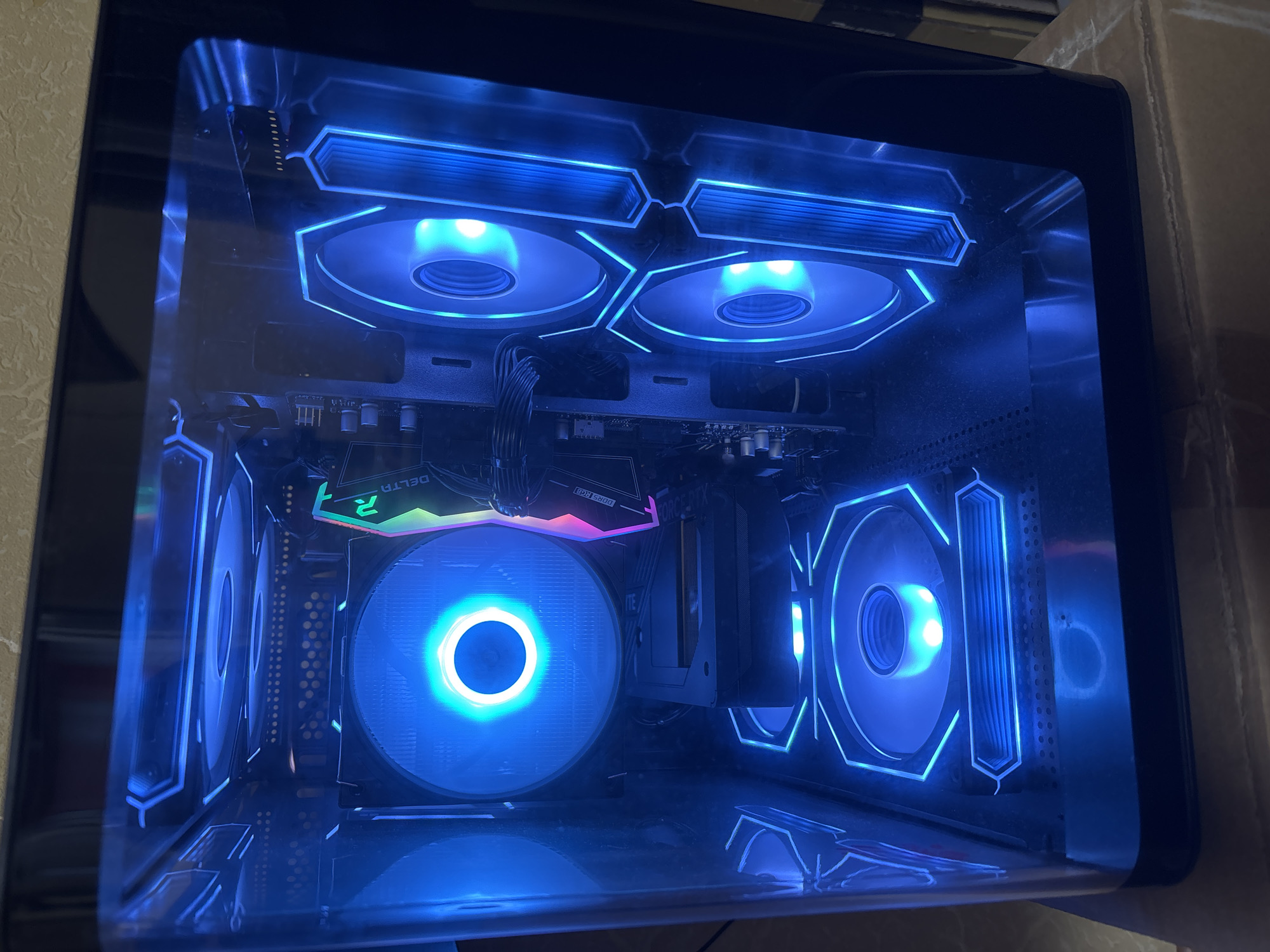 A high-angle, full-shot shows a computer tower with blue RGB lighting, viewed from the side through a transparent panel. The computer is placed on top of cardboard boxes, suggesting it is in storage or has recently been delivered. The computer tower itself is illuminated with bright blue lights from multiple fans and accent lighting, creating a futuristic aesthetic. The internal components are visible, including a large CPU cooler with its own RGB ring, a graphics card with "RTX" and "3080" branding, and RAM modules. The overall scene is dark, with the computer's lighting being the primary light source. There are no people or animals present in the image. The background is dark and out of focus, obscuring any details of the surrounding environment. The image is a close-up of the computer and its lighting, with no discernible elements that would indicate its location in Naberezhnye Chelny, Russia.