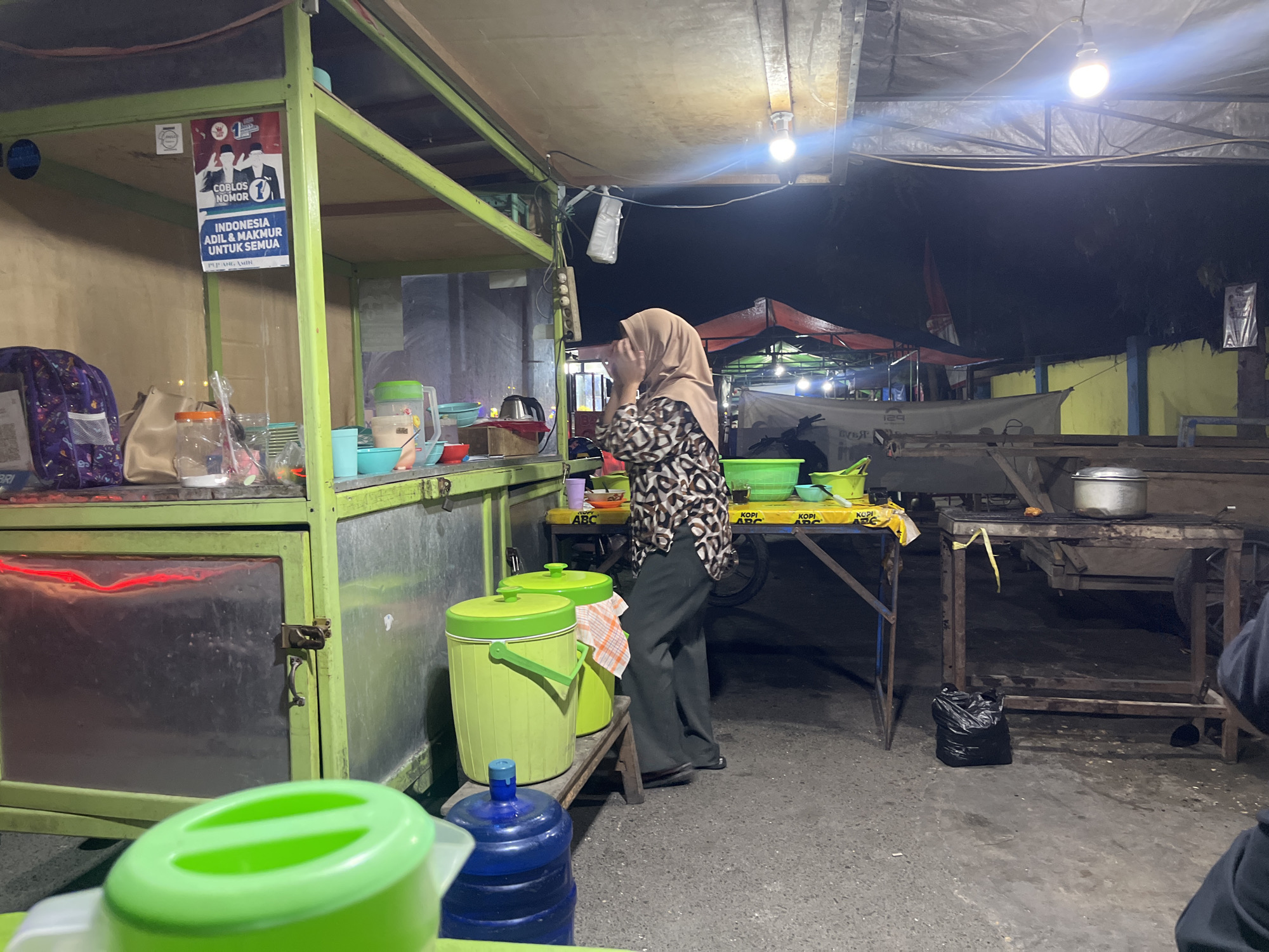 In Luwuk, Indonesia, an outdoor food stall operates at night under a tarp and string lights. A person wearing a hijab and patterned shirt stands near a serving counter, facing away from the camera. To the left, a green cart displays a poster with an election slogan and images of two men. Various containers and items are visible on the cart, including buckets, plastic bottles, and bags. A small, open cooking area with a grill and pots is situated to the right. The ground is paved, and a black trash bag lies on the pavement. The scene suggests a typical street food vendor setting.