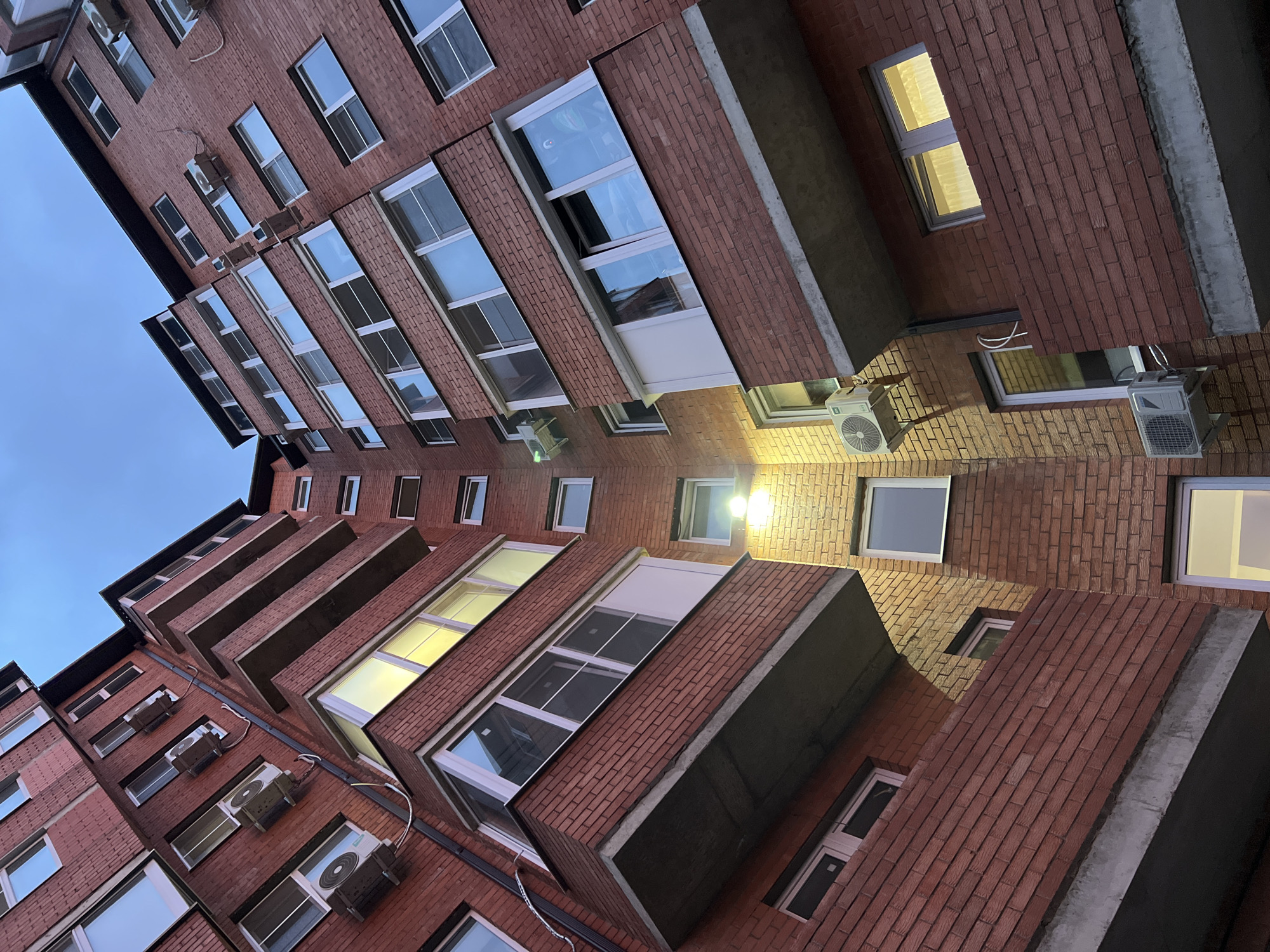 The image shows a low-angle view of a brick building in Yablonovskiy, Russia. The architecture is modern, featuring multiple floors with protruding balconies or sections, creating a stepped facade. The building has numerous windows, some reflecting the sky and others illuminated from within. Air conditioning units are mounted on the exterior walls. The sky visible at the top left corner appears to be dusk or dawn, with a mix of blue and gray hues. The overall impression is urban and residential.