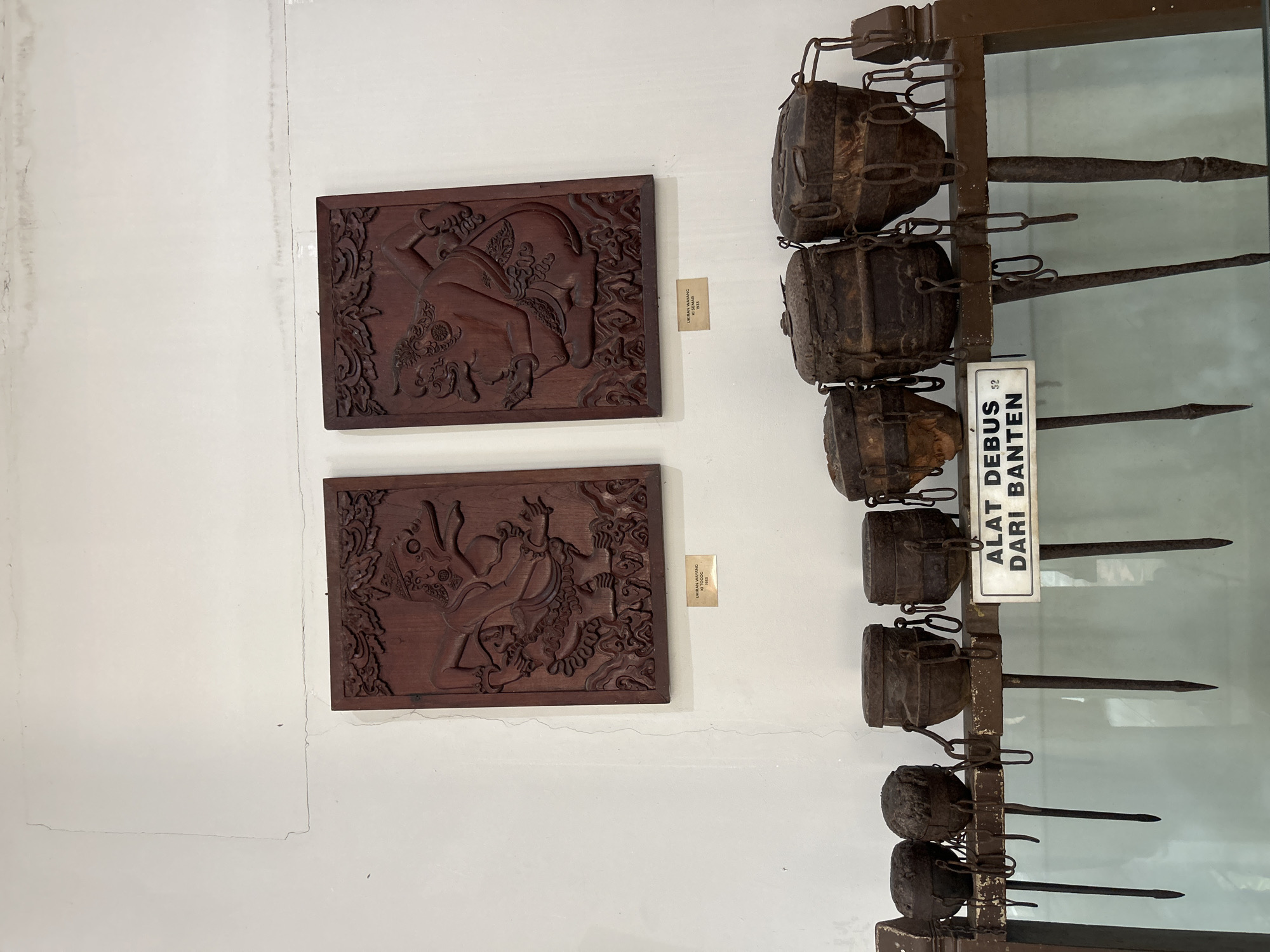 The image depicts a display within a museum, likely the Museum Pusaka Keraton Kasepuhan in Cirebon, Indonesia, given the presence of Wayang reliefs and traditional tools. Two wooden reliefs of Wayang figures, possibly characters from Javanese puppetry, are mounted on a white wall. Below these reliefs, a collection of what appear to be historical ritualistic or ceremonial tools is displayed on a wooden shelf behind glass. These tools consist of several cylindrical, metal containers with chain attachments, each suspended by a long, pointed metal stake. A white sign with black text in Indonesian, "ALAT DEBUS DARI BANTEN," is placed in front of the tools, identifying them as "Debus tools from Banten." The overall setting suggests an indoor exhibition space with neutral-colored walls and a display cabinet.