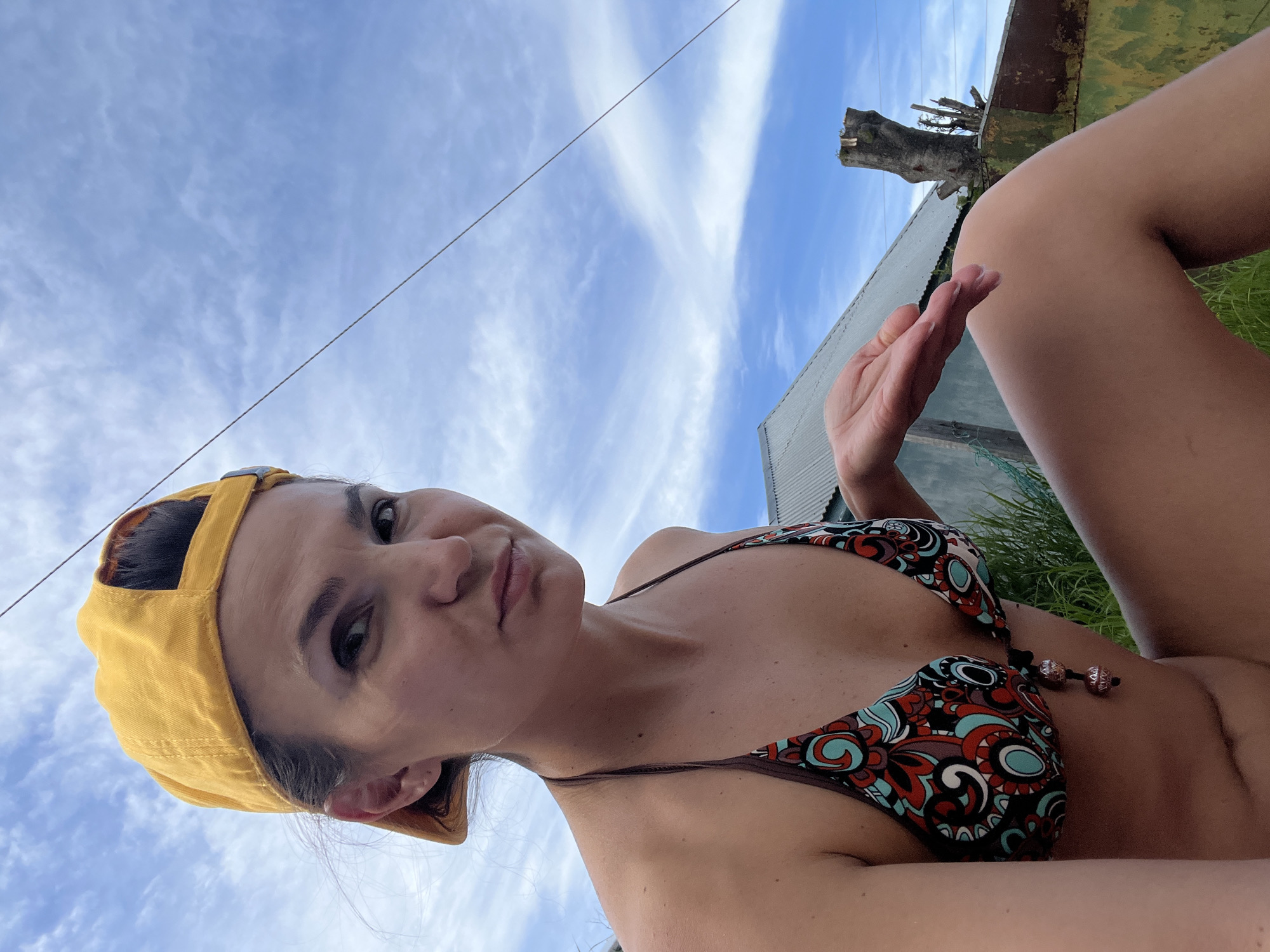 A woman wearing a yellow baseball cap and a patterned bikini is photographed from the chest up against a blue sky with wispy clouds. Her head is tilted, and she looks directly at the camera with a slight frown. Her left arm is bent at the elbow and extended forward, with her hand open. In the background, a corrugated metal roof of a building and a dead tree stump are visible. The scene appears to be outdoors, possibly in a rural or semi-rural setting within or near Petropavlovsk-Kamchatsky, Russia, suggested by the typical architecture and vegetation.