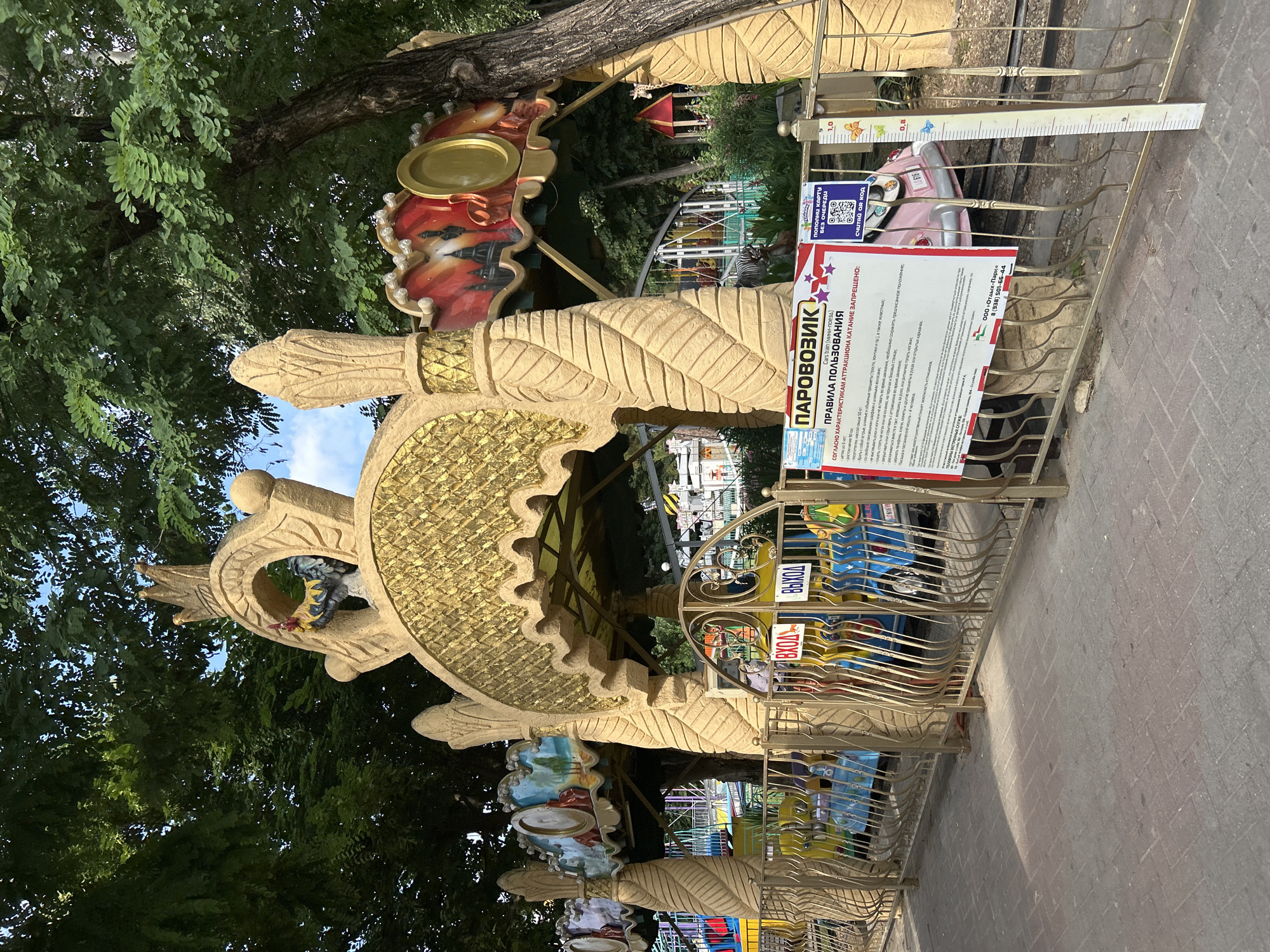 The image shows a children's carousel ride in the city of Gelendzhik, Russia. The carousel has a train-themed design with colorful carriages and a decorative golden archway featuring a crown and a rooster. A sign in Russian provides rules and regulations for the ride. The carousel is surrounded by a metal fence, and the ground is paved with bricks. Trees with lush green foliage form a backdrop, and the sky is visible through the branches. There are no people clearly visible in the image.
