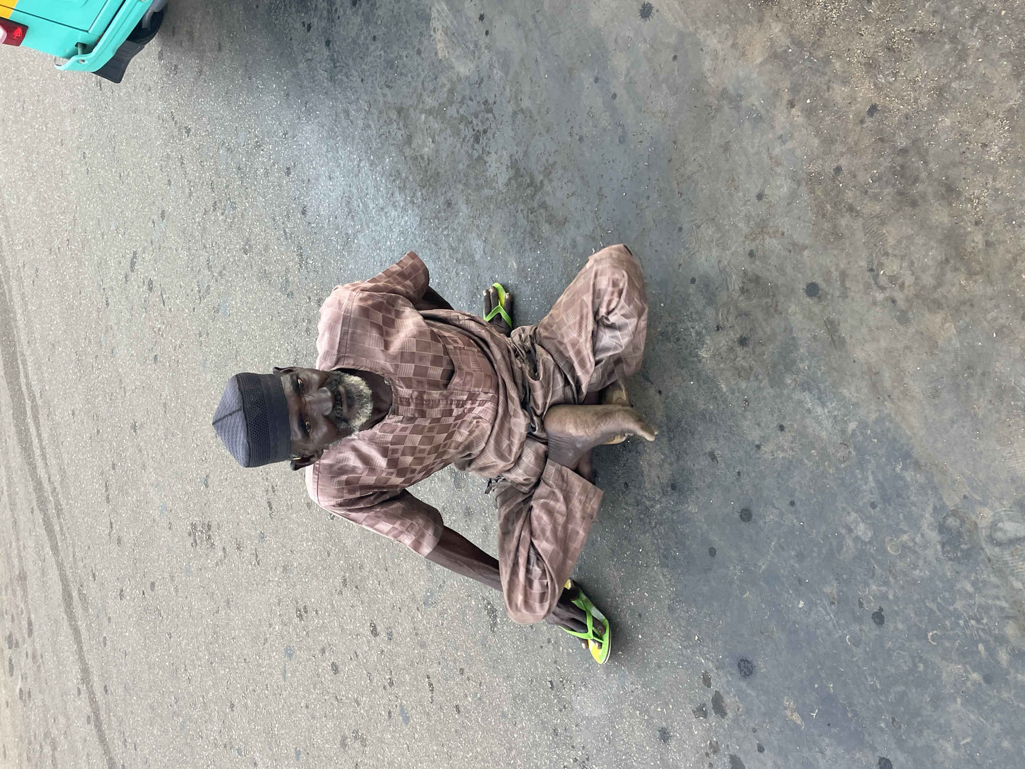An elderly man, wearing a light brown patterned dashiki and a black cap, is sitting on the ground with his legs crossed in a street setting. He has a grey beard and is looking directly at the camera. His left foot is adorned with a bright green flip-flop. To the right of the frame, a portion of a turquoise and yellow vehicle is visible, suggesting an urban environment. The ground is asphalt, showing signs of wear and tear with scattered dark spots and some lighter debris. The lighting suggests an overcast day.