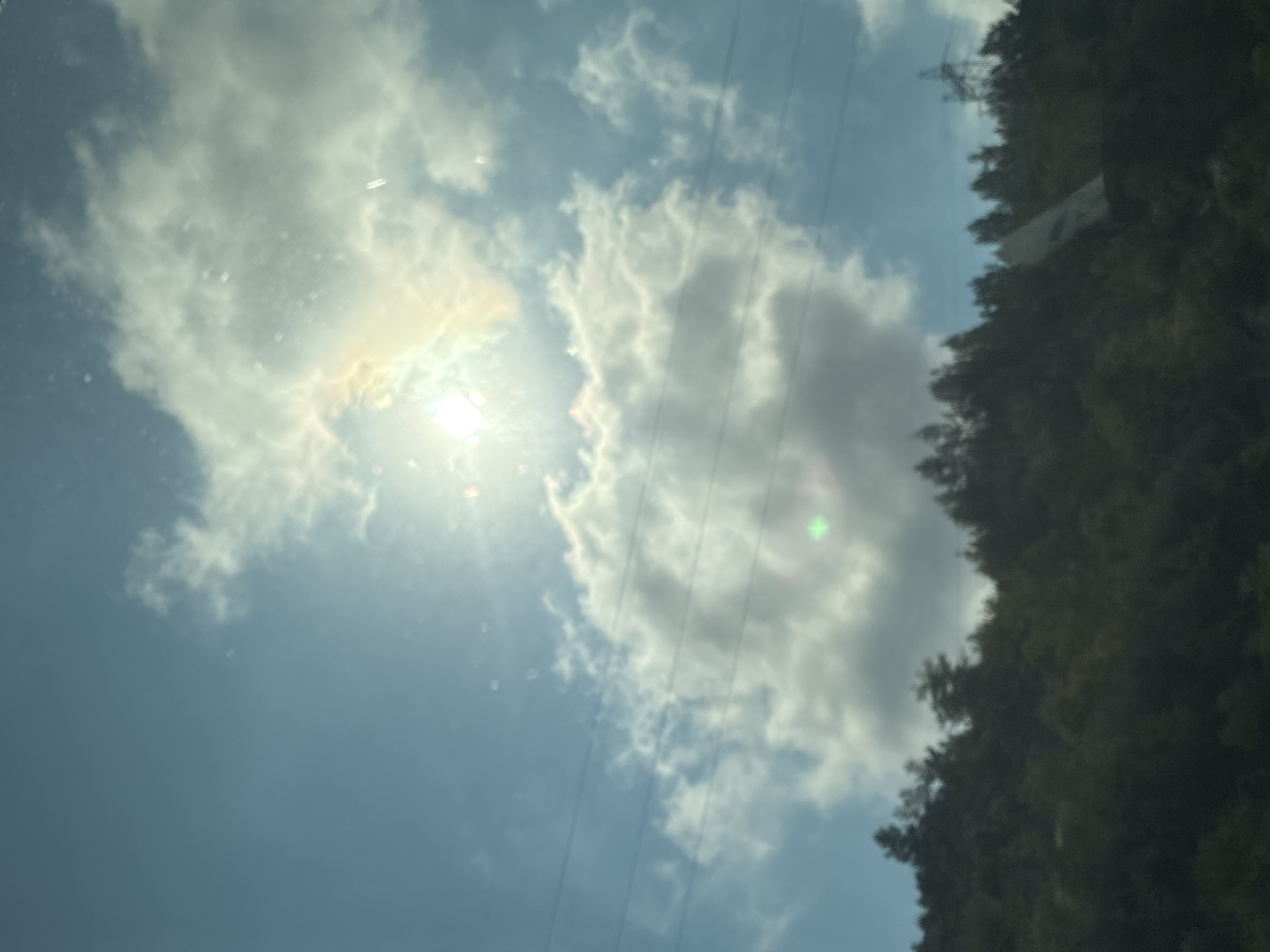 The image depicts a sunlit sky with scattered clouds above a densely wooded hillside. The sun is bright and partially obscured by clouds, casting a warm glow. Several power lines are visible crisscrossing the sky. The hillside is covered with dark green trees, and in the distance, a power pylon can be seen on the right side of the frame. The image appears to be taken from a slightly elevated perspective, possibly from a vehicle, looking towards the hillside and the sky. No people are visible in the image. There are no specific identifiable landmarks to indicate the city of Alanya, Turkey. The image is slightly out of focus and exhibits lens flare.