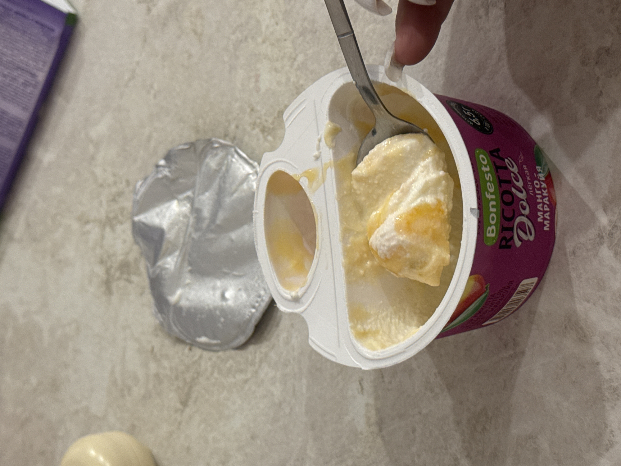 In Saratov, Russia, on a grey speckled countertop, a half-eaten container of Bonfesto Ricotta Dolce yogurt with mango and passion fruit flavors is displayed. A spoon is partially submerged in the creamy yogurt, scooping a portion. The yogurt container is purple with text and fruit imagery, and the foil lid is peeled back and lies behind the container. To the left of the yogurt, a pale yellow, spherical object is visible on the countertop. In the background, a portion of a purple and green package with text is also visible. A person's hand with manicured nails is partially visible on the right side of the image, interacting with the spoon.