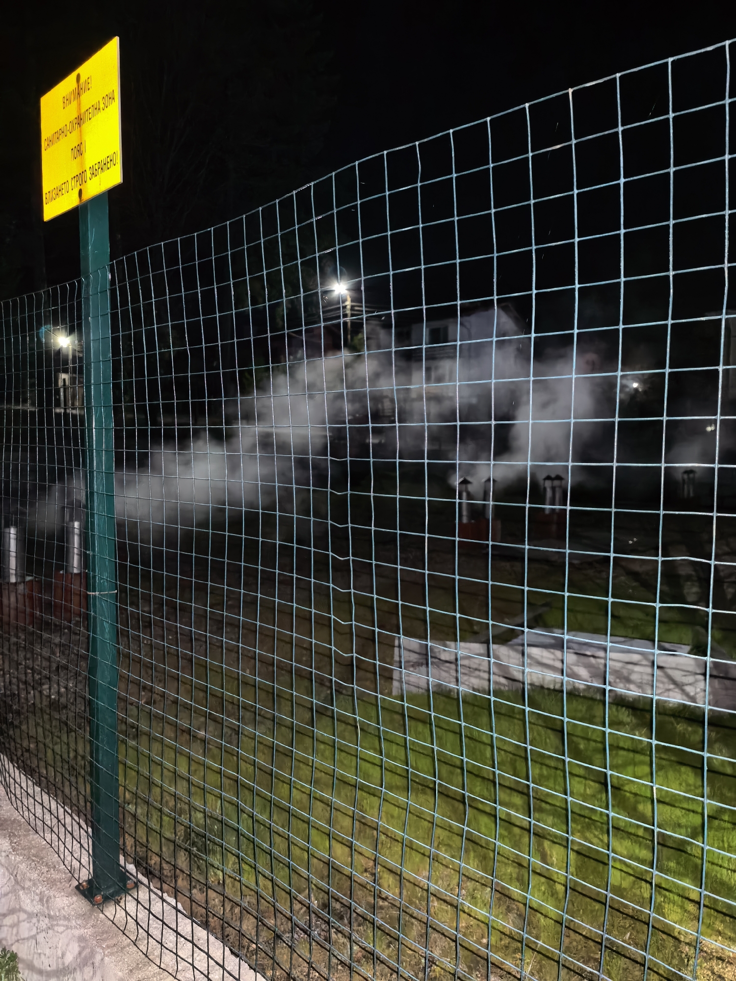 At night in an unknown city, a green chain-link fence stands between a sidewalk and an area emitting steam from several chimneys. A yellow sign with Cyrillic text, mounted on a green pole, is visible next to the fence. Streetlights illuminate the scene.