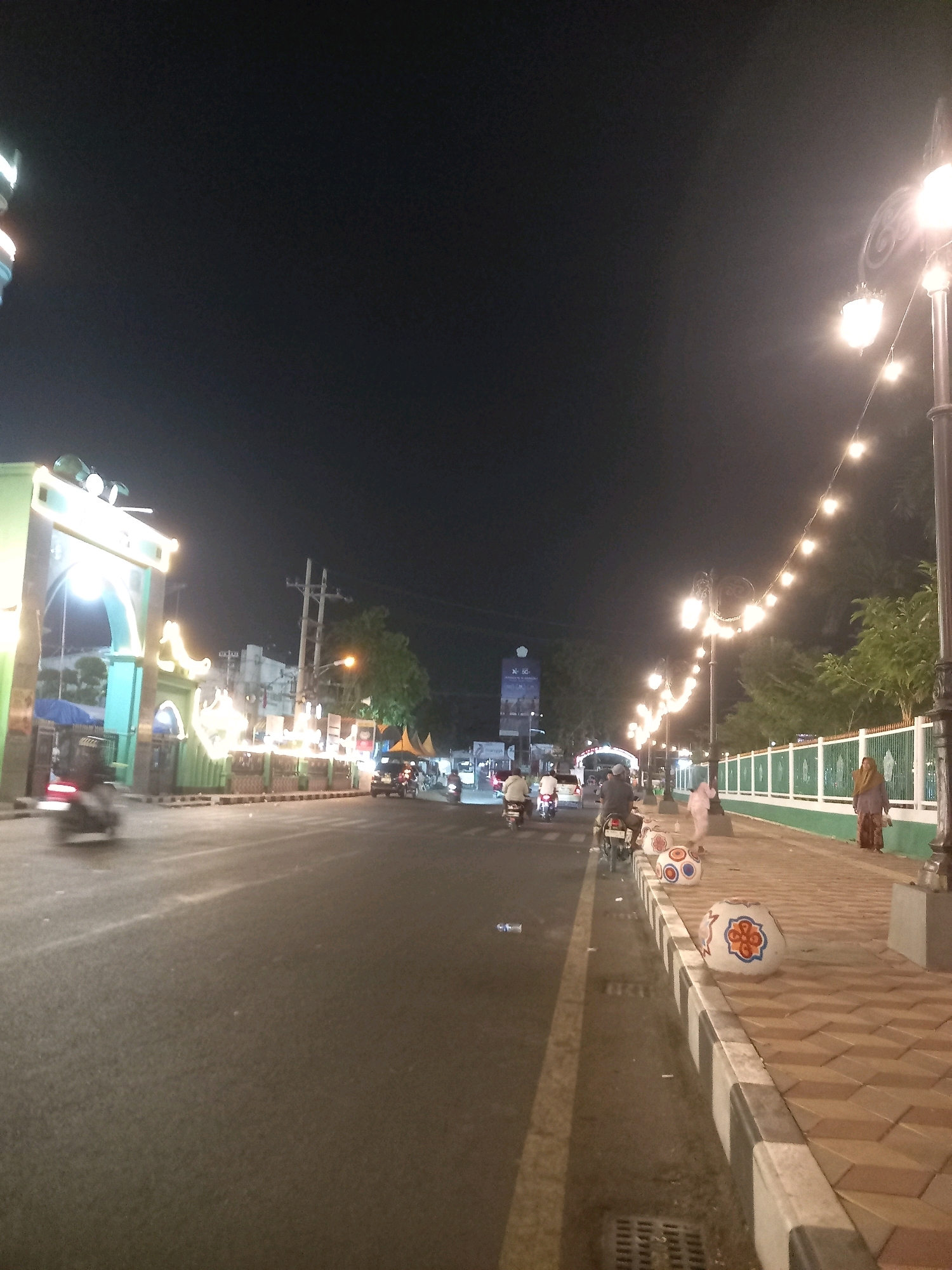 The image depicts a street scene at night in Pamekasan, Indonesia. The foreground shows a paved road with a raised sidewalk on the right. The sidewalk is decorated with several large, spherical objects featuring colorful patterns. A white fence runs along the edge of the sidewalk, separating it from a green area behind. Streetlights with strings of small lights are visible overhead, illuminating the scene.
Several people are present. On the sidewalk, two women are standing, and another person is walking. On the road, multiple motorcycles are being ridden by people, and one car is visible. On the left side of the image, there's a large, brightly lit archway, possibly an entrance to an area or an event. Buildings and trees are visible in the background, along with utility poles and wires. The overall atmosphere is that of a lively street at night.