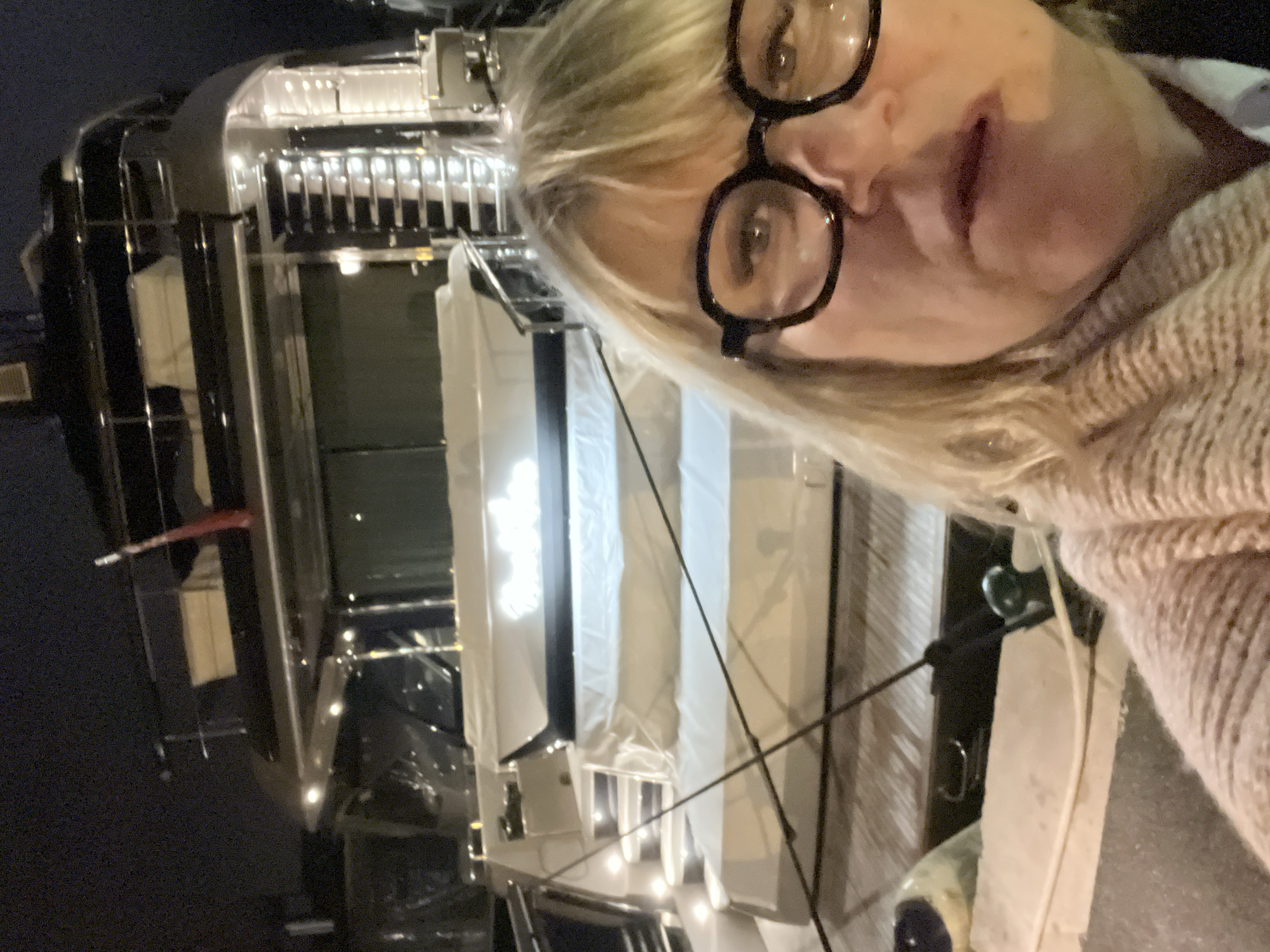 A woman with blonde hair and glasses stands on a dock next to a large, dark-colored yacht at night. The yacht is illuminated with bright lights on the deck and stairs. The scene is likely at a marina in Grimaud, France, as indicated in the prompt. The woman is taking a selfie. Ropes connect the yacht to the dock.