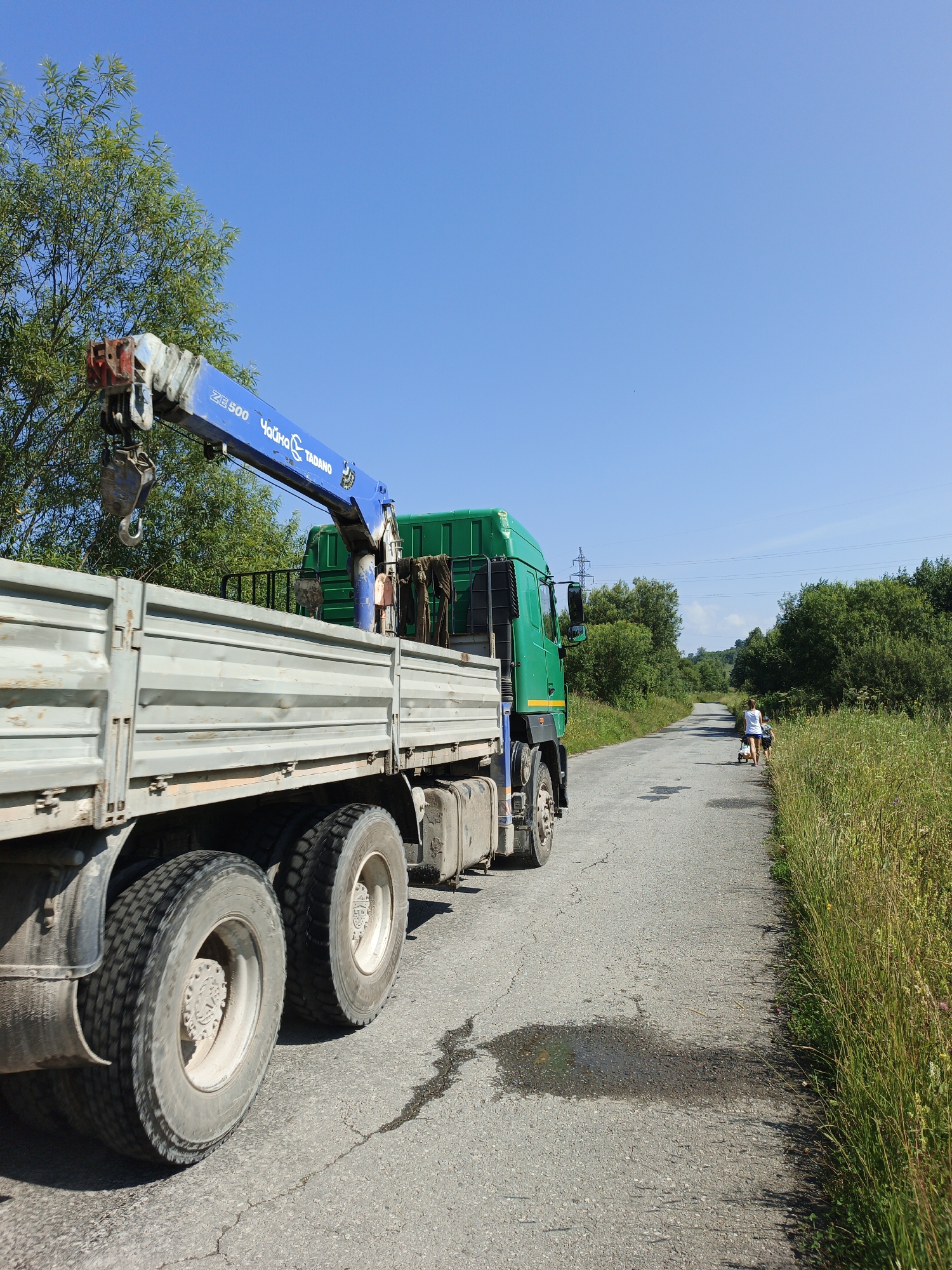 A large, green truck with a blue crane attachment is parked on the side of a paved rural road. The truck's trailer is grey and appears to be empty. The road is narrow and flanked by tall, green grass and trees. In the distance, two people, one appearing to be an adult and the other a child, are walking away from the camera down the road. The sky is clear and blue. The scene suggests a sunny day in a rural setting. The location of Urtiushka, Russia, is not visually discernible from the image itself.