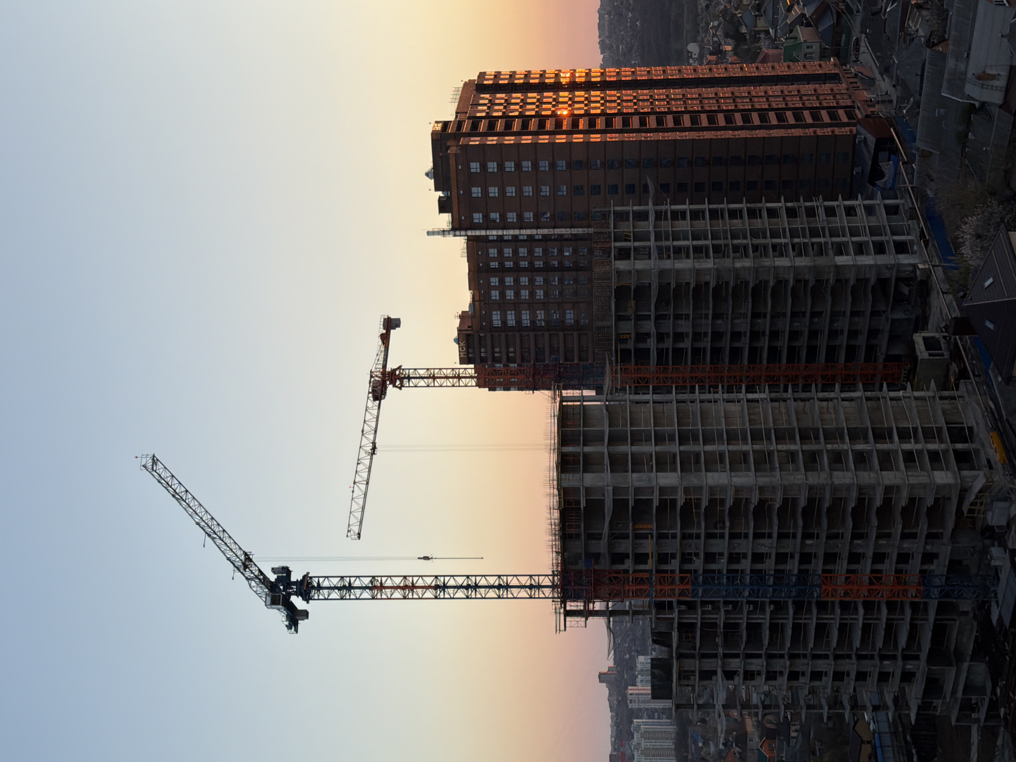 The image shows a construction site in an urban environment, likely in the city of Azov, Russia. Two high-rise buildings are visible, one partially completed with exposed concrete framework and another, taller building with a brown facade and visible windows. Three cranes are positioned around the construction site. The sky has a gradient of color ranging from light blue to orange, suggesting sunrise or sunset. No people are directly visible in the frame. The buildings are surrounded by lower-rise urban development, visible in the background.