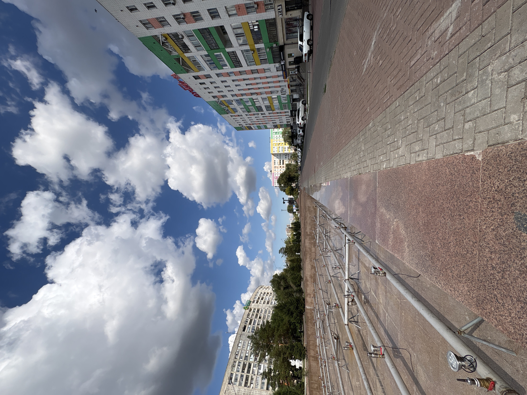 The image captures a street scene in Pavlodar, Kazakhstan, likely taken on a sunny day with scattered clouds. In the foreground, an empty fountain structure with metallic pipes and nozzles is visible. To the right of the fountain, a paved walkway made of reddish-brown bricks and grey cobblestones runs alongside a street. The street is lined with several multi-story apartment buildings, characterized by their colorful facades, featuring sections of white, green, yellow, and pink. Cars are parked along the street and on the sidewalk. Further down the street, more apartment buildings and some trees are visible, suggesting an urban environment. The sky is a bright blue, with large, fluffy white clouds dominating the upper portion of the image. The overall impression is one of a well-maintained urban public space.
