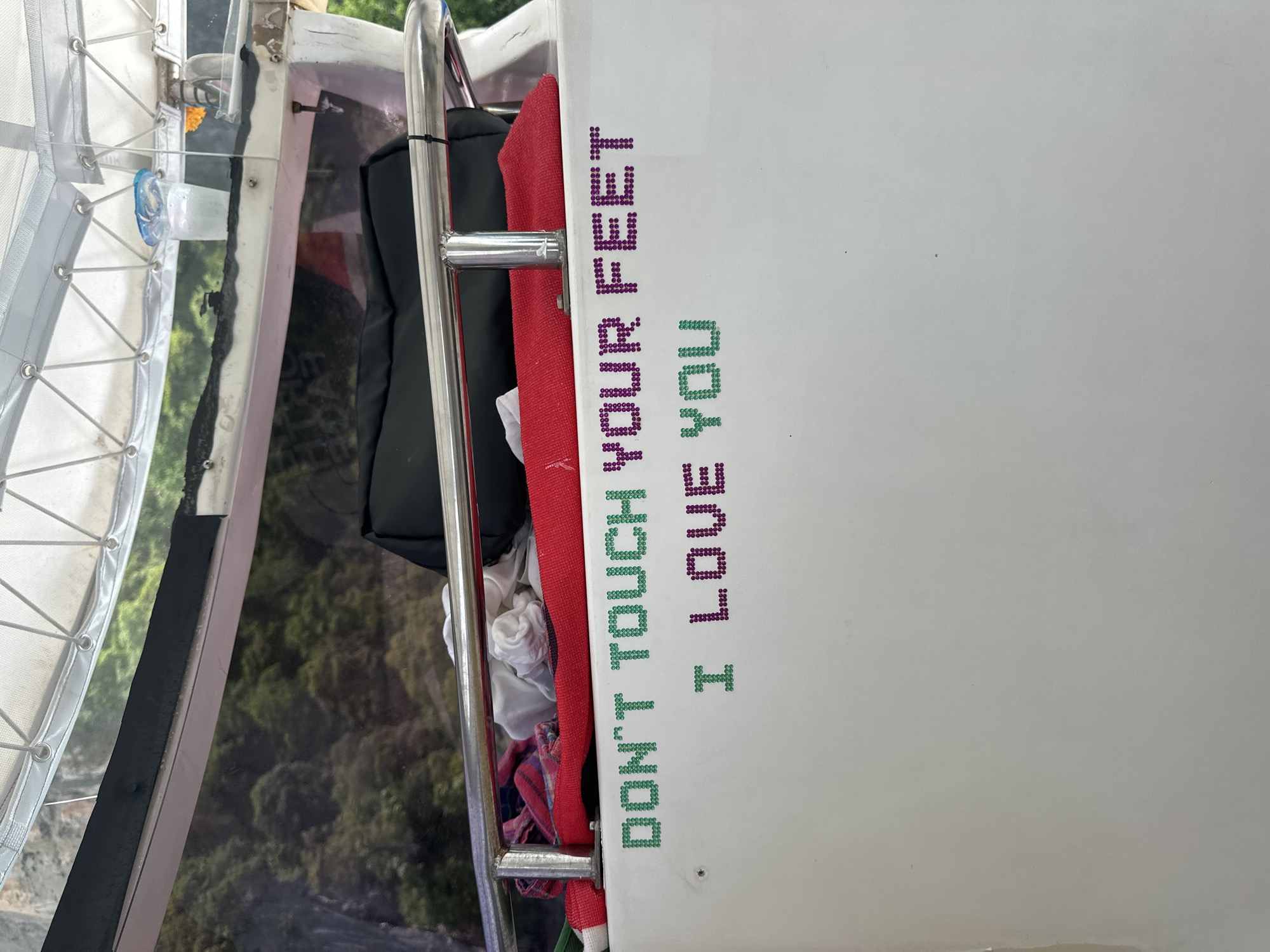 The image shows a white boat with a metal railing. Attached to the boat's railing are several pieces of clothing, including a red cloth and some white fabric. Below the railing, on the white surface of the boat, are two lines of text made of colorful, beaded letters. The top line reads "DON'T TOUCH YOUR FEET" in green and purple letters. The bottom line reads "I LOVE YOU" in green and purple letters. The background of the image is a blurred landscape of green foliage and rocky cliffs, suggesting the boat is in a natural, possibly tropical, setting. The scene is likely located at Haad Hin Khom, Thailand, given the context of the image.