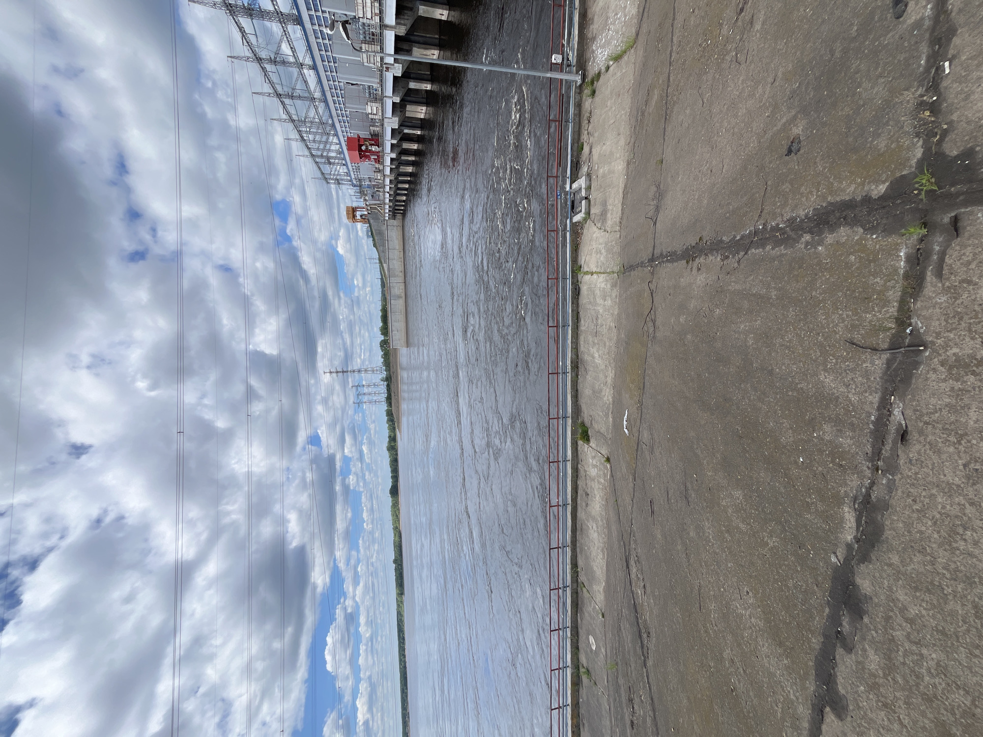The image captures a view of the Votkinsk Hydroelectric Power Station's spillway on the Kama River, located in Chaikovsky, Russia. The sky is filled with a mix of white and gray clouds against a blue backdrop. Several power lines stretch across the sky, supported by tall pylons in the distance.
The foreground consists of a concrete embankment, cracked and showing signs of wear, with sparse vegetation growing in the cracks. A red railing runs along the edge of the embankment overlooking the river.
The Kama River is wide and appears to be flowing rapidly, with turbulent water and swirling patterns indicating a strong current. The water's color is a murky brown.
On the right side of the image is the impressive structure of the hydroelectric power station. It features multiple levels with white and gray buildings, supported by large concrete pillars that extend into the river. Red and orange structures, likely part of the power generation equipment, are visible on the station's roof. Power lines connect to tall transmission towers situated on the station's upper levels.
In the far distance, the opposite bank of the river is lined with trees and vegetation. The overall scene depicts a large industrial infrastructure integrated with a powerful natural watercourse under a dynamic sky.