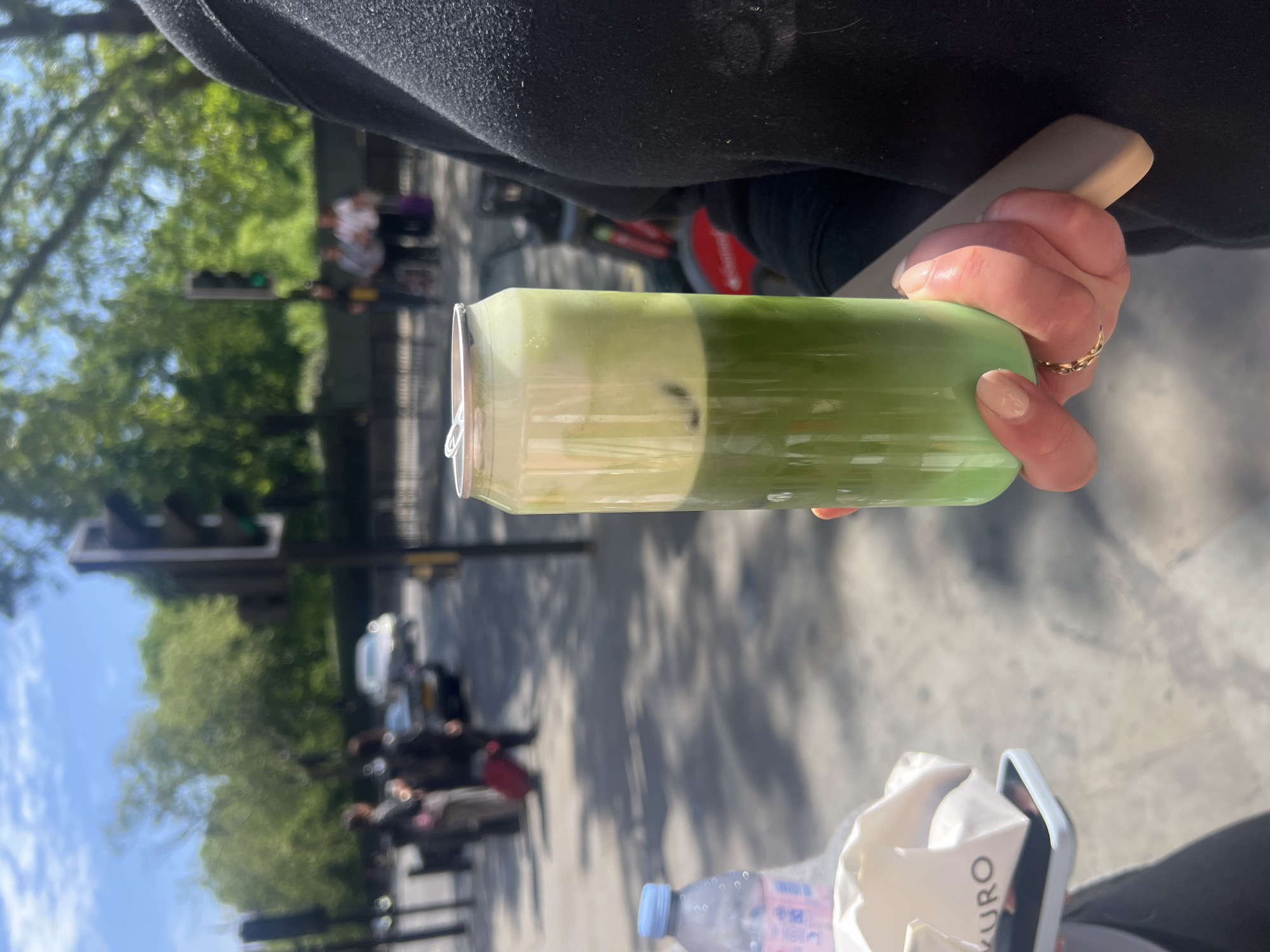 The image shows a person holding a can-shaped glass of a green drink, likely matcha latte, in their right hand. The person is wearing a black sweater and a gold ring on their ring finger. Below the drink, there is a water bottle and a white box with the word "KURO" printed on it. In the background, there is a street scene with trees, a traffic light, people sitting, and a parked car. Based on these details, the location could be in London, United Kingdom.