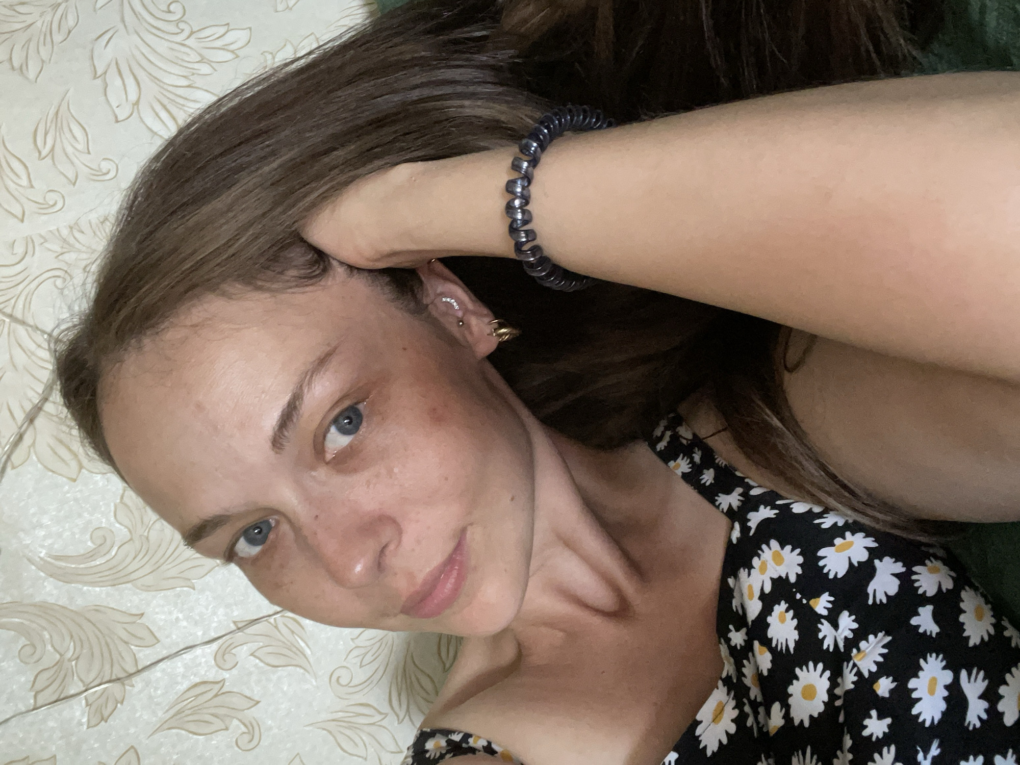 A young woman with brown hair and blue eyes, wearing a black and white floral-print dress, is pictured lying down. She has a bracelet on her arm and piercings in her ears. The background is a patterned wallpaper. The image appears to be a selfie taken indoors.