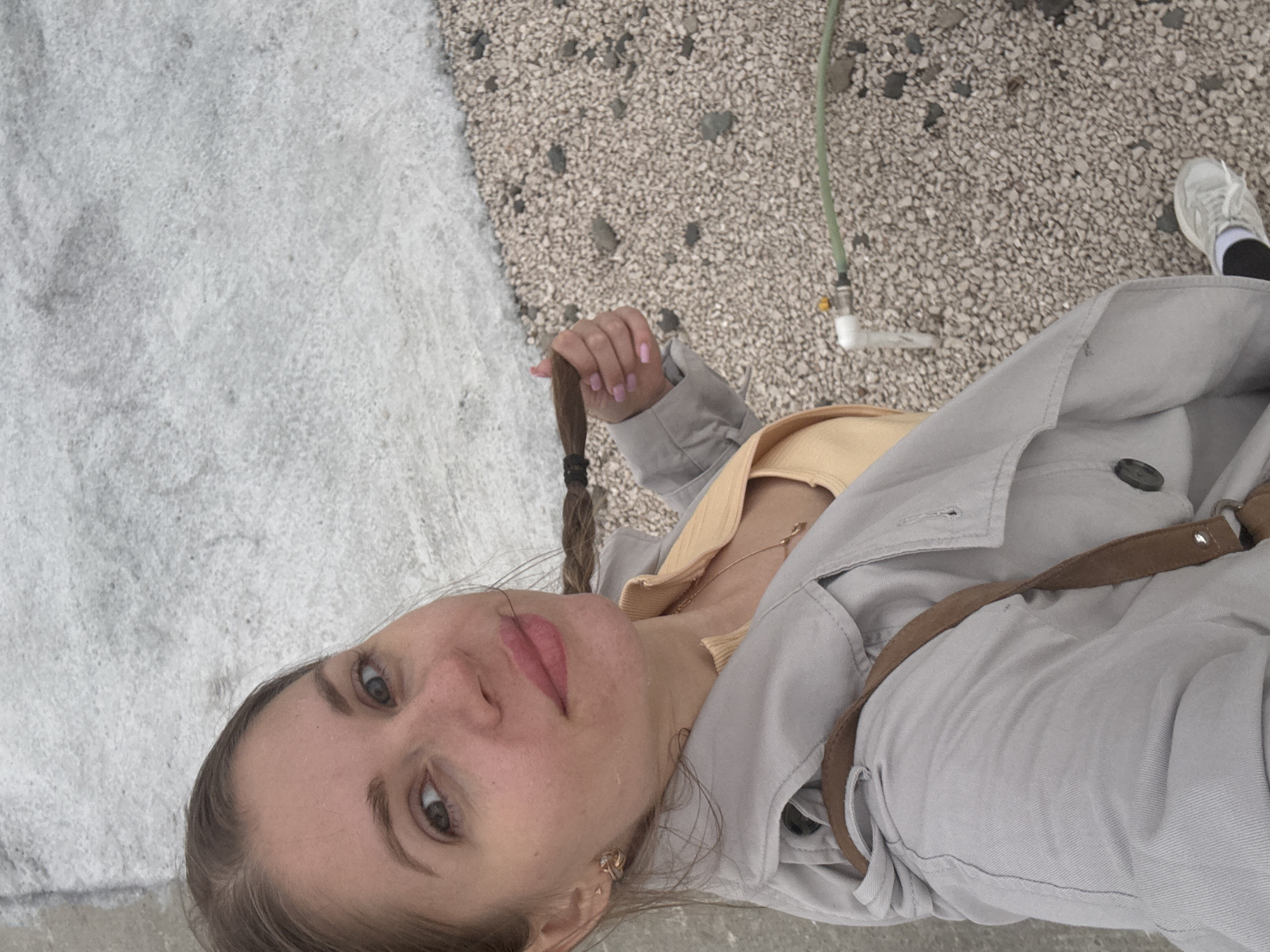A person is seen from a low angle, looking toward the camera, with a background showing two distinct textures. To the left, a large, rough, white surface suggests either snow or a highly textured light-colored material. To the right, a patch of gray pebbles or small stones provides another textural contrast. A green hose and a white fixture extend from the pebble area. The person has light skin, brown hair styled in a braid held at the end, and is wearing a yellow top and a gray coat with a brown strap across the torso. A single white sneaker is visible at the top right. The location appears to be a coastal area based on the pebbles; however, the specific location in Batumi, Georgia, is impossible to determine from the given image.